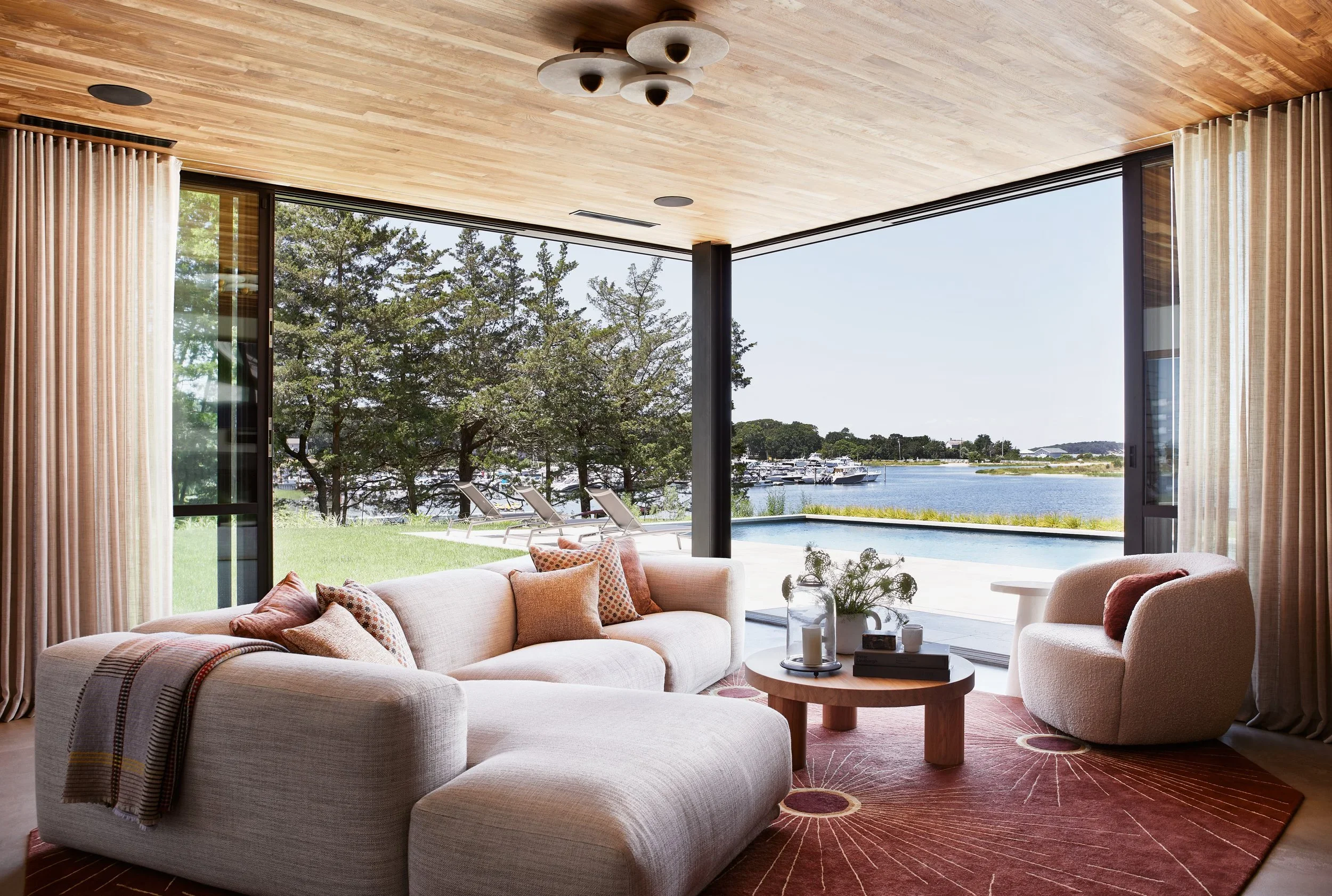 Living room with large glass windows overlooking a lake and outdoor pool, decorated with a beige sectional sofa with pillows, a round coffee table with flowers and candles, and an armchair, with a red rug underneath.