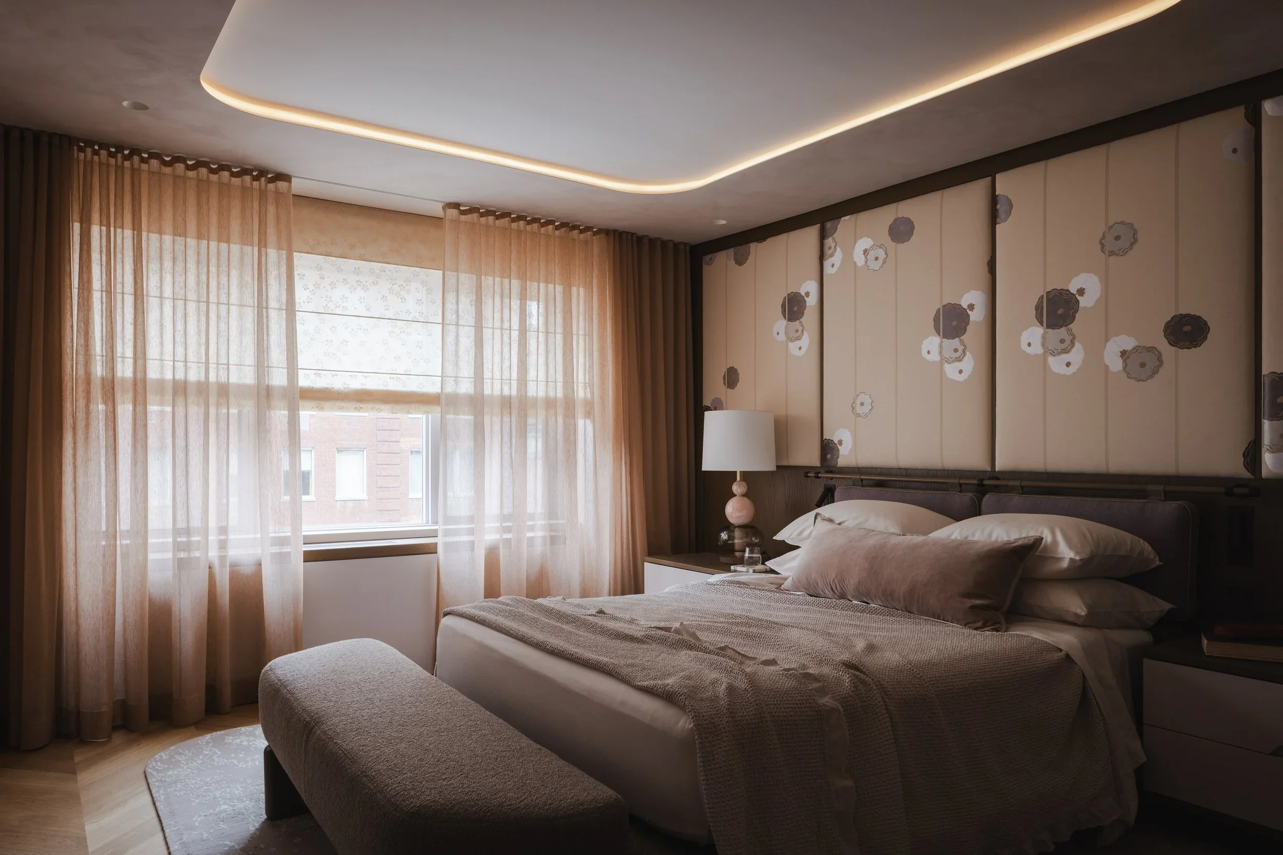 A bedroom with a bed, pillows, side table with a lamp, beige curtains, a window showing a brick building outside, and decorative wall panels with a floral pattern.