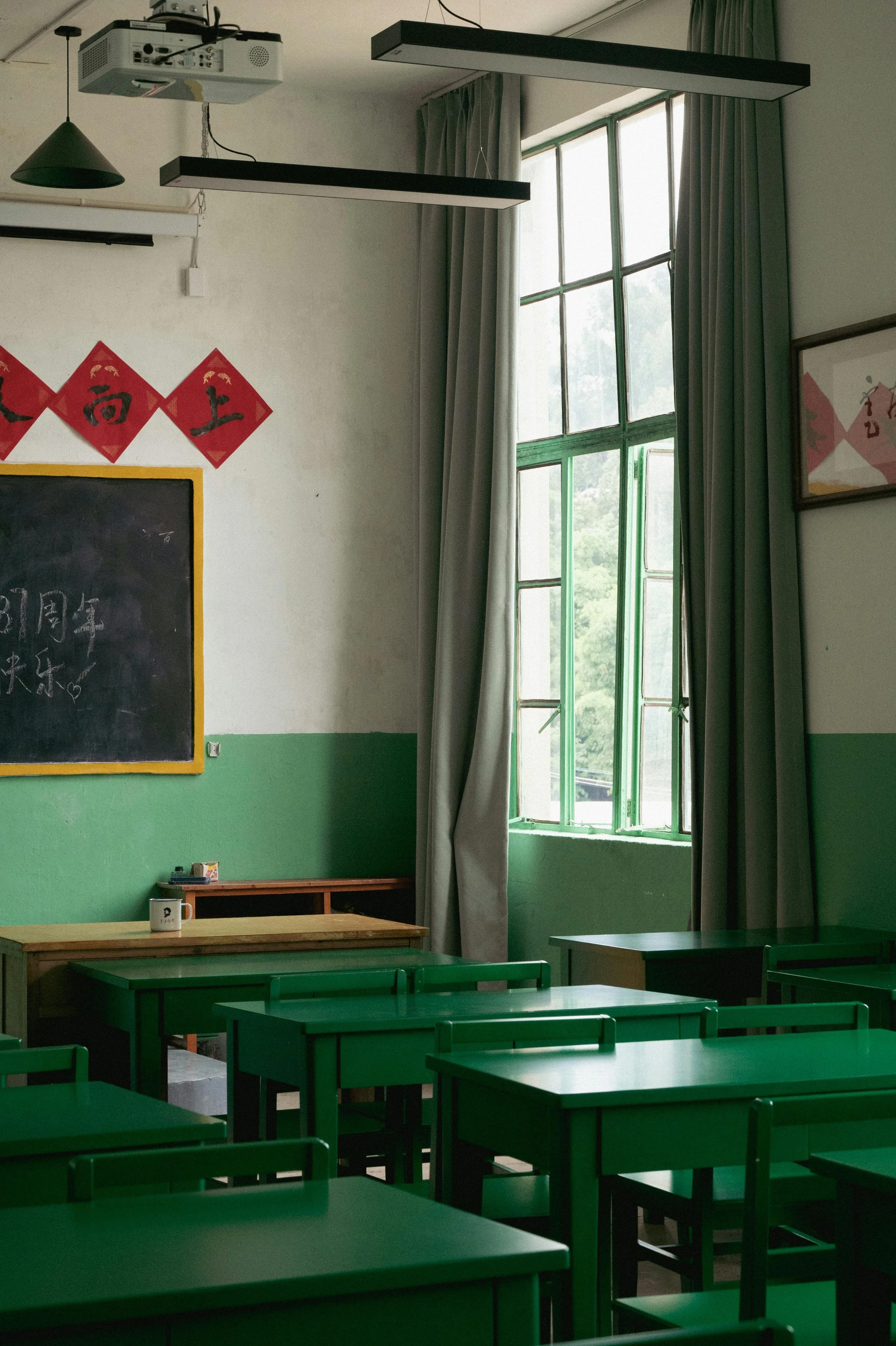 An empty classroom with green desks and chairs, a chalkboard with Chinese writing, red Chinese banners on the wall, and large windows with curtains letting in natural light.
