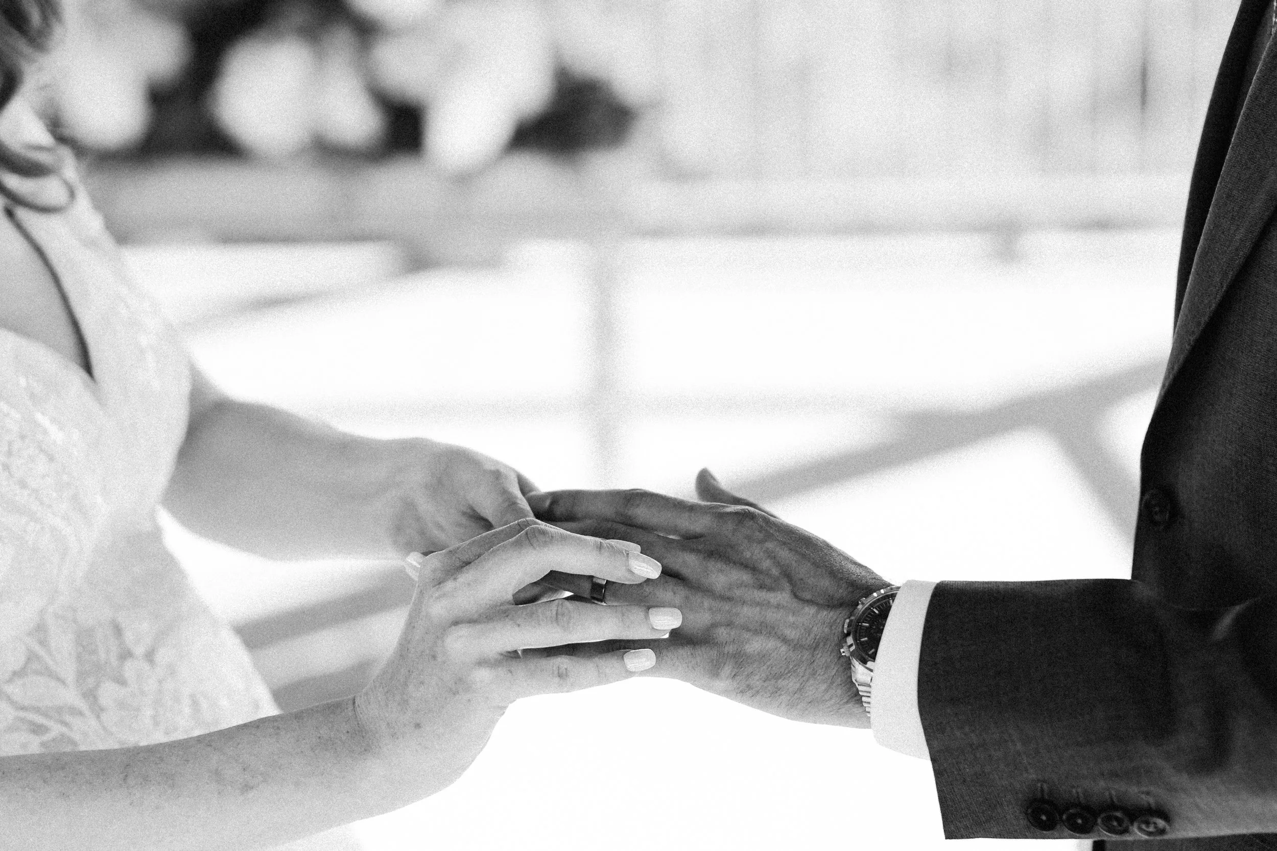 A black and white photo of a couple holding hands during a wedding ceremony. The woman is wearing a wedding dress and the man is in a suit.