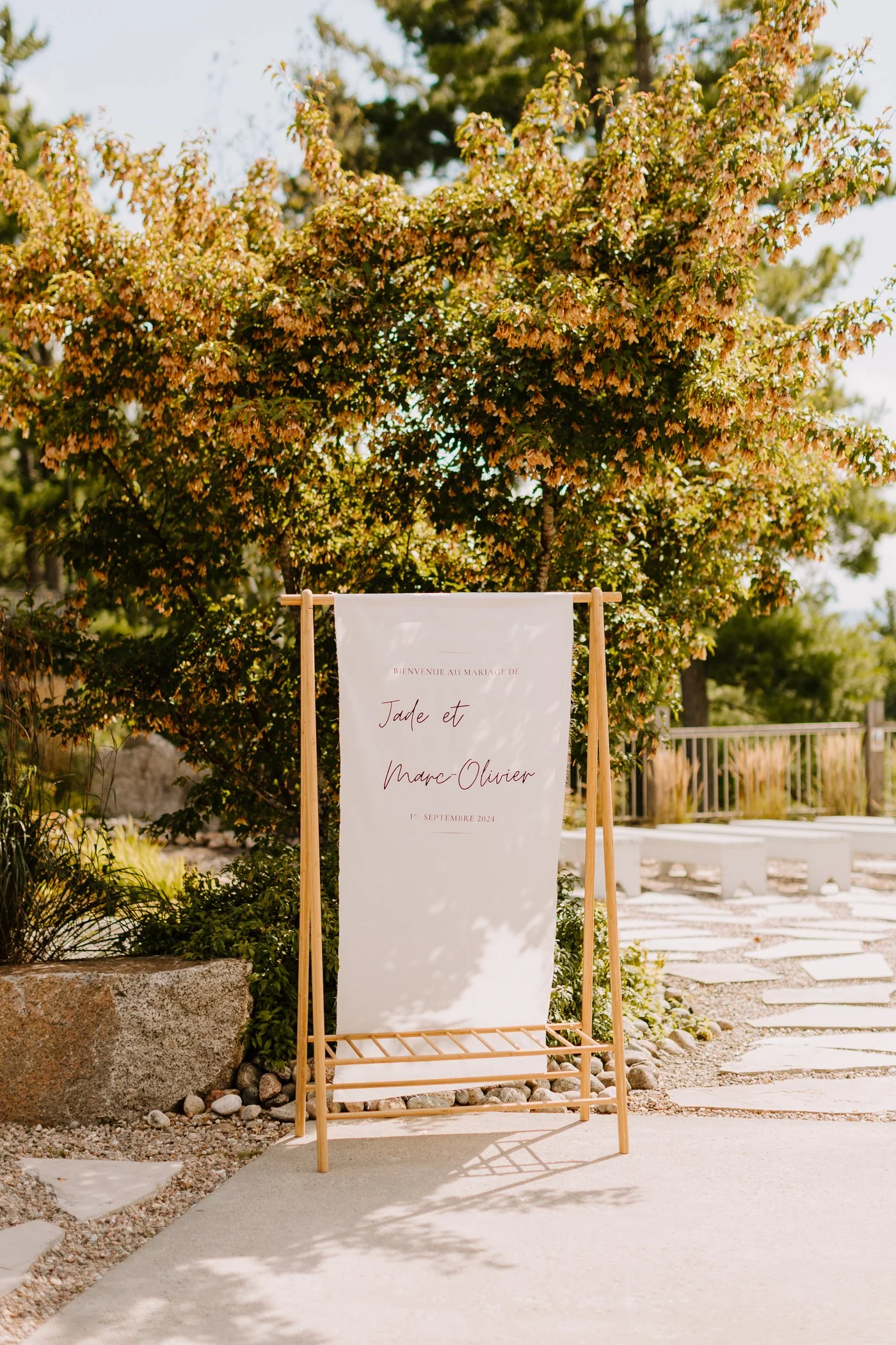 A white fabric wedding or event sign with elegant red and black script, supported by a wooden easel, situated outdoors with trees and a stone pathway in the background.