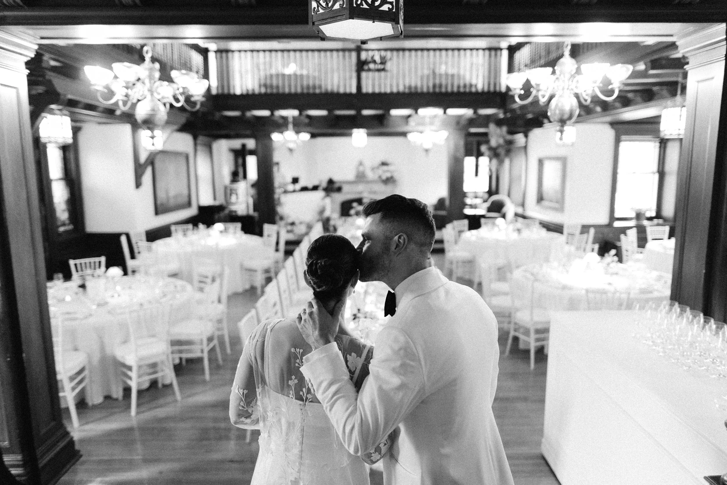 A black and white photograph of a couple dancing closely in a decorated banquet hall, seen through a mirror. The hall features chandeliers, framed artwork, and neatly arranged tables with white chairs.