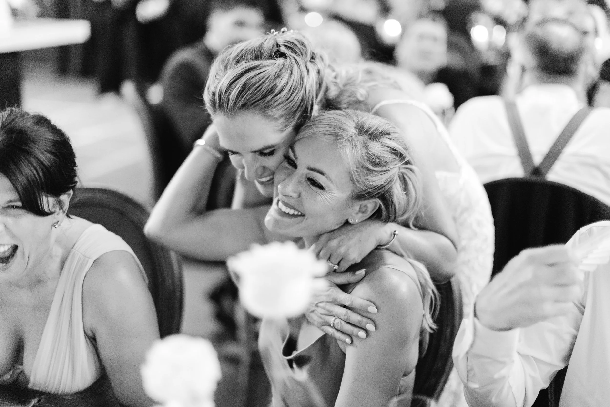 Two women sharing an embrace at a social gathering, both smiling, in a black-and-white photo.