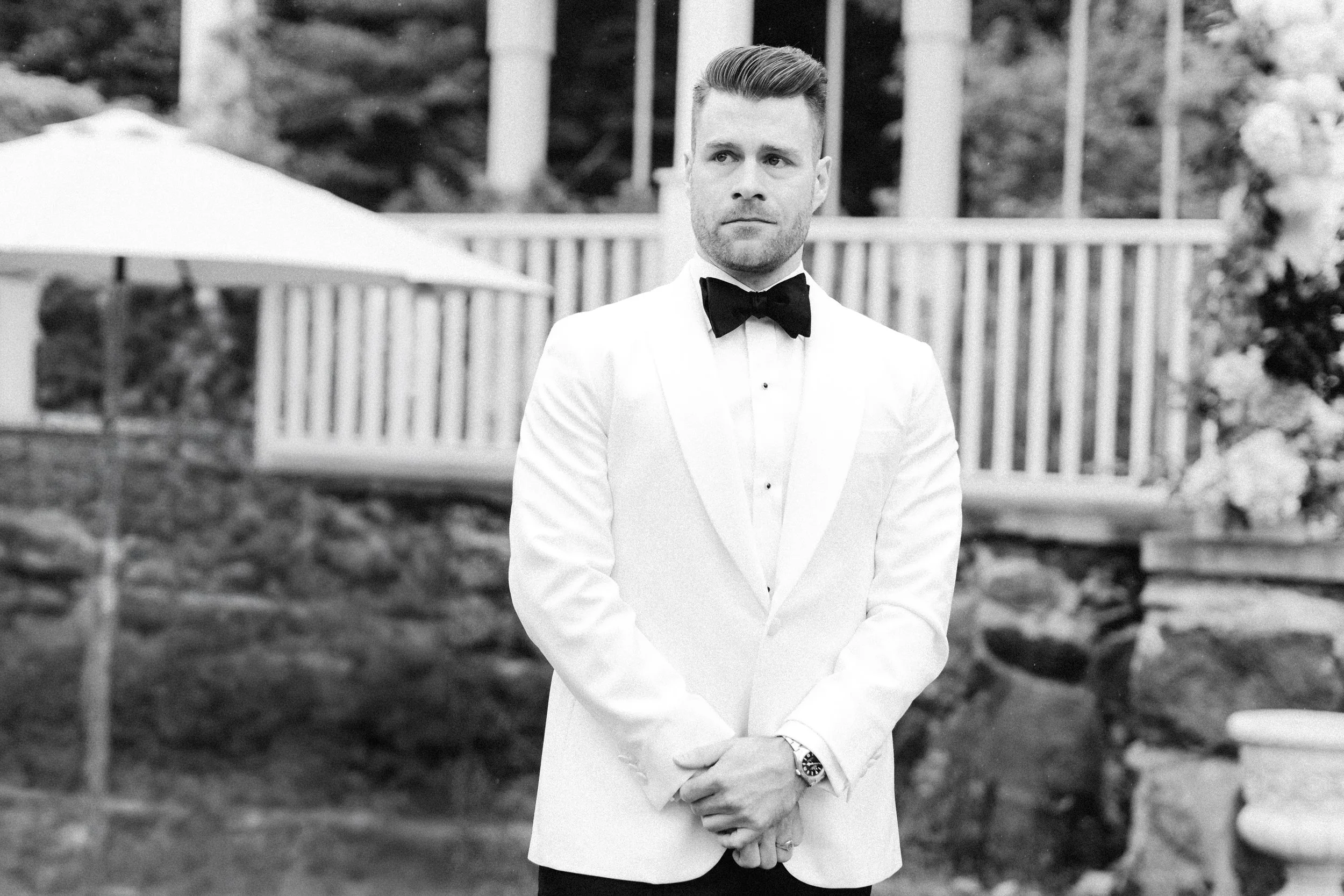 A man dressed in formal attire, wearing a white tuxedo with a black bow tie, standing outdoors in front of a white fence and some decorative elements, captured in black and white.