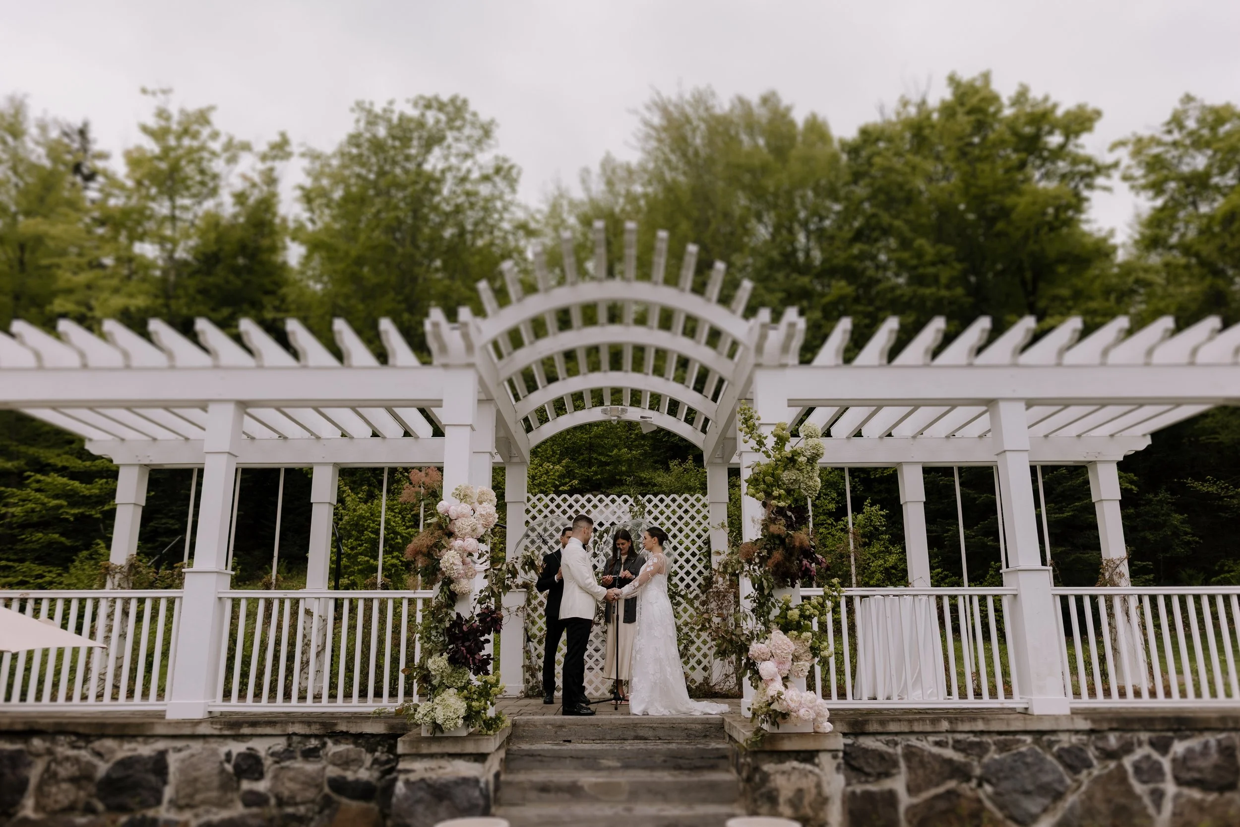 A couple getting married at an outdoor wedding ceremony under a white pergola with floral decorations, surrounded by green trees on a cloudy day.