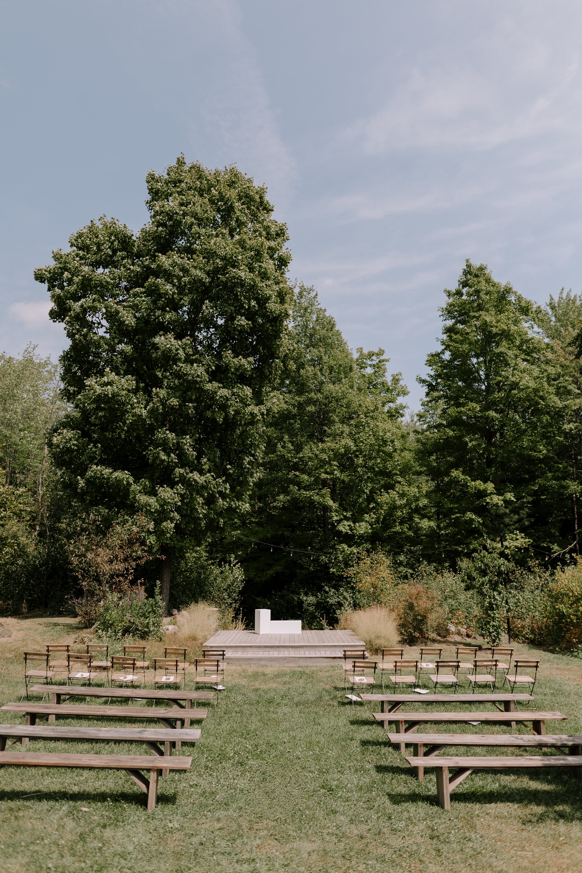 Outdoor outdoor wedding ceremony setup with chairs facing a stage and woodland backdrop under a blue sky.