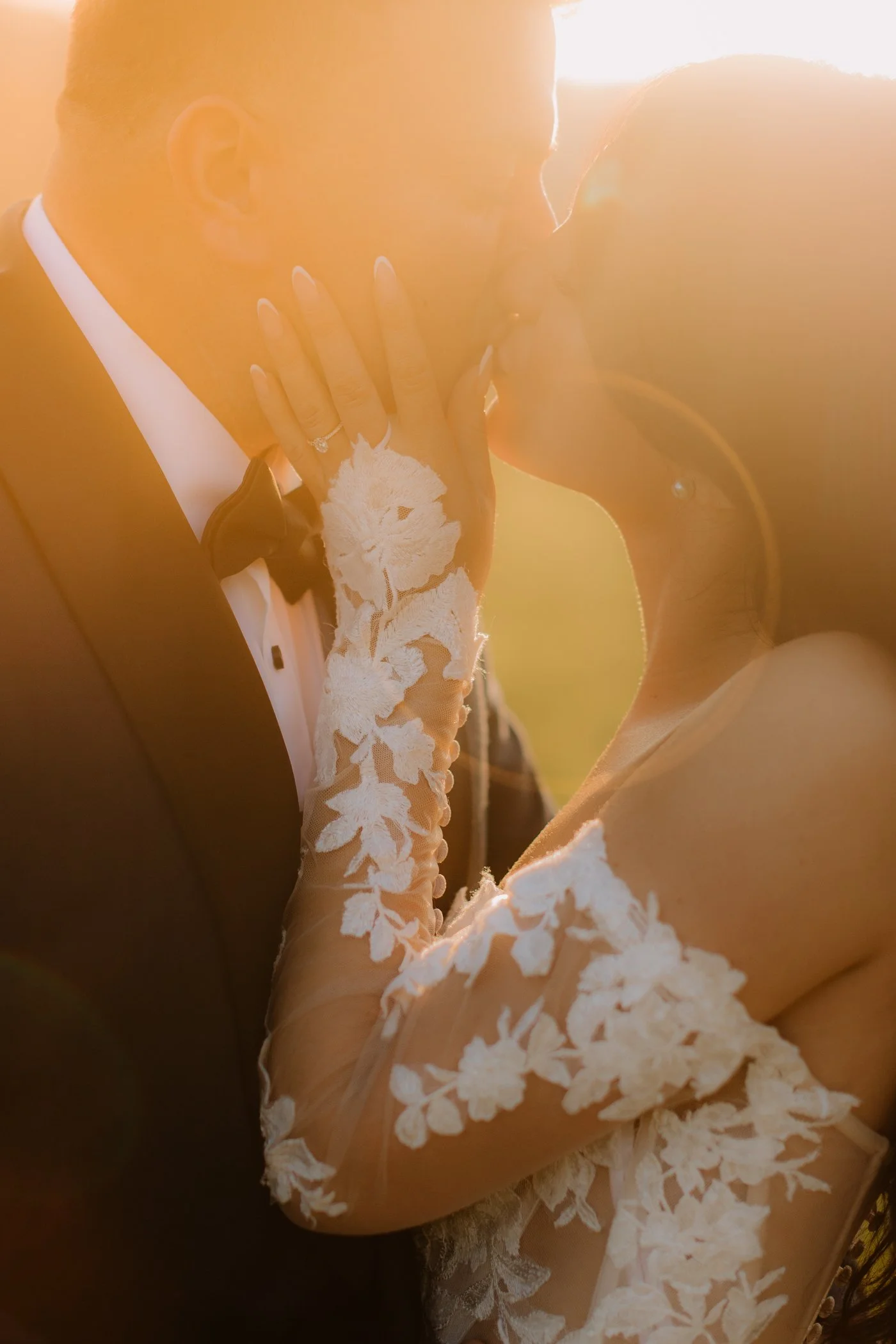 A bride and groom sharing a kiss, with the bride's hand on the groom's face, in front of bright sunlight, both dressed in wedding attire.
