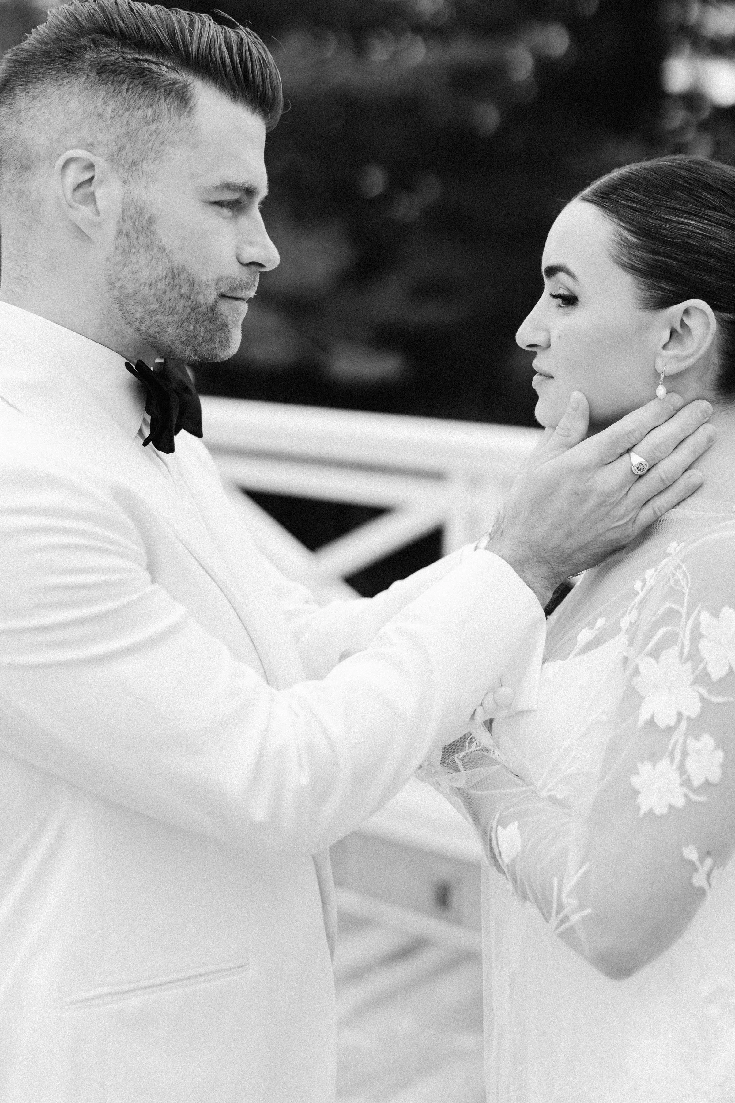 A man and woman holding each other's faces, gazing into each other's eyes, in an intimate moment, dressed in formal attire, black and white photograph.