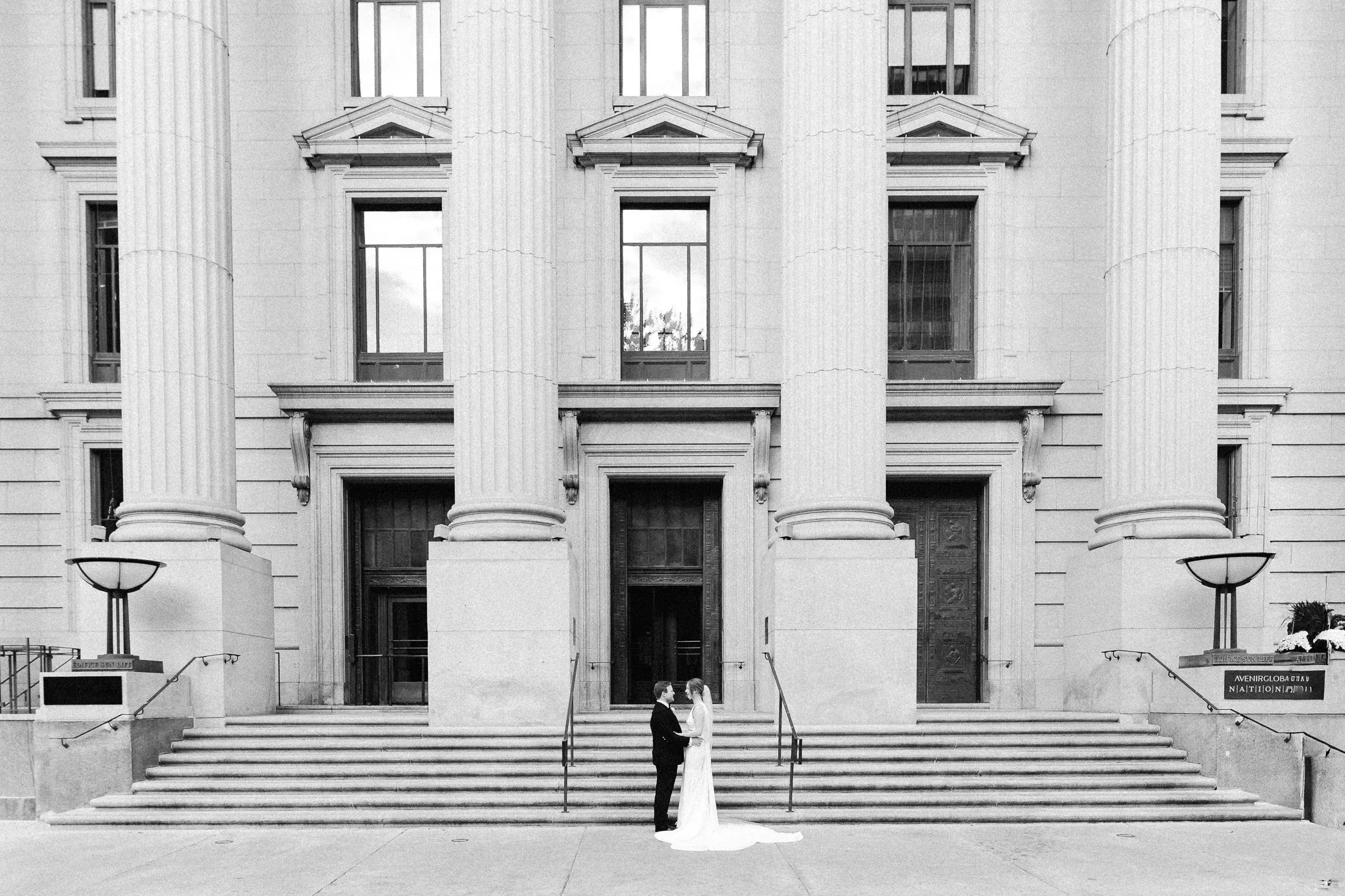 A black and white photograph of a couple standing on the stairs of a classical-style building with large columns, possibly during a wedding or special event.