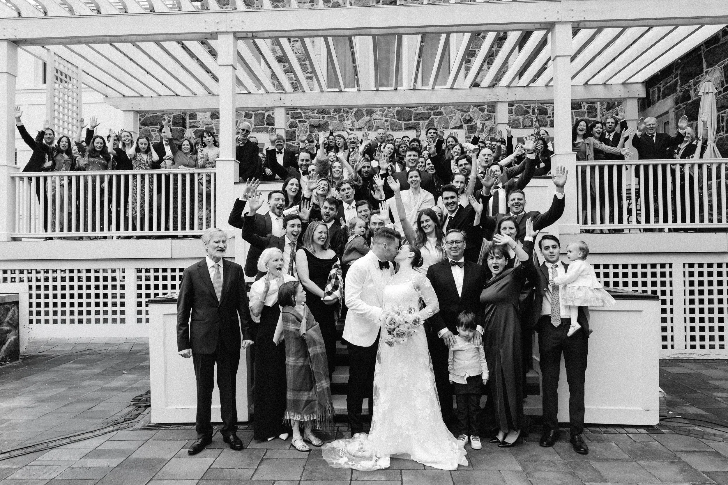 A large group of people dressed in formal attire gathered outdoors, with some raising their hands and smiling. In the front, a bride in a wedding dress and a groom in a tuxedo are standing together, surrounded by family and friends. The setting appea