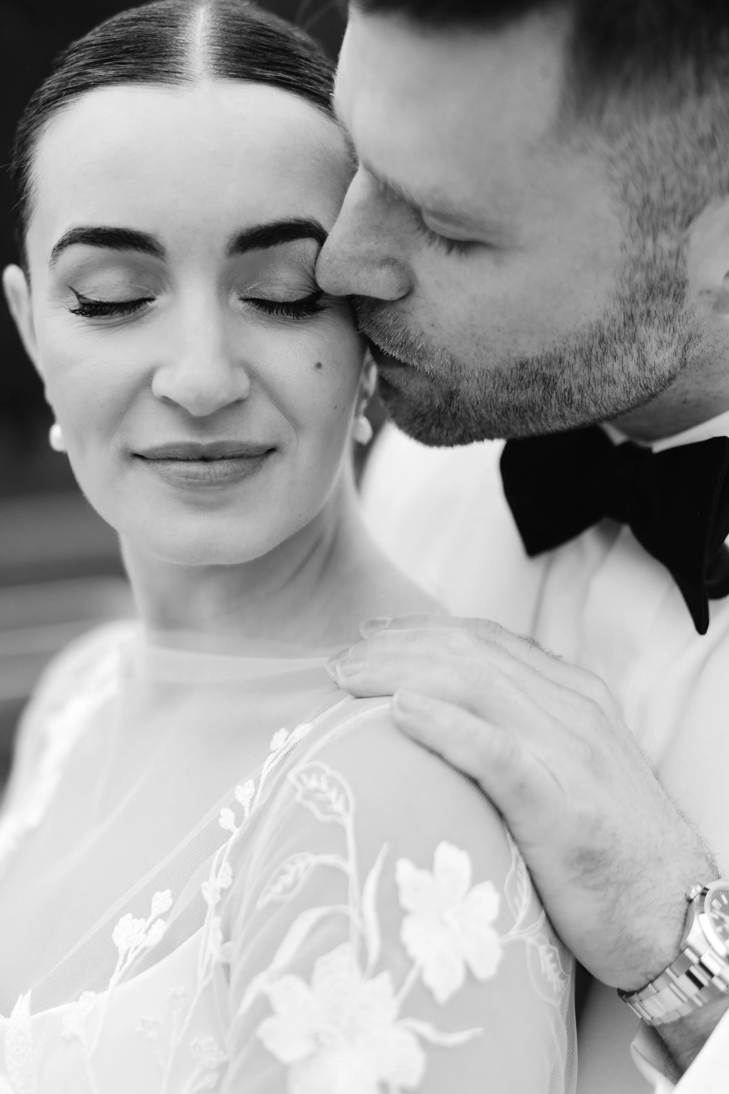 A grayscale close-up photo of a woman with closed eyes, smiling slightly, and a man kissing her on the forehead. The woman is wearing a dress with floral embroidery. The man has short hair, a beard, and is wearing a suit with a bow tie, and a watch o