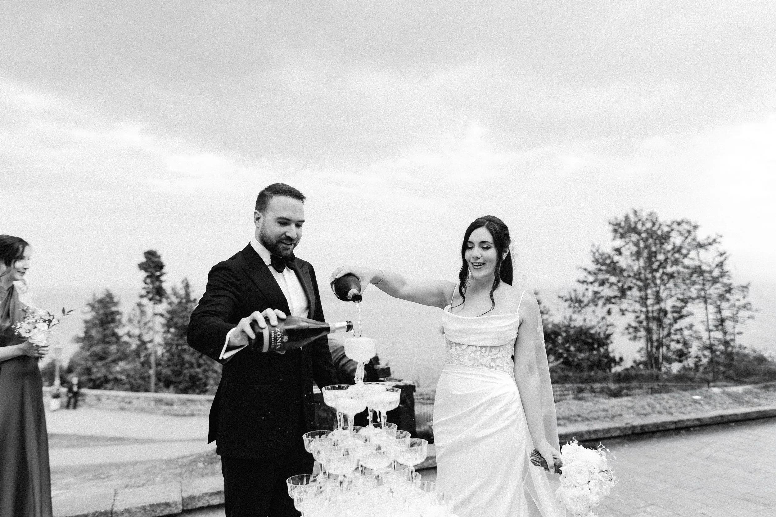 A groom and bride pouring champagne into a multi-tiered cake at an outdoor wedding reception in the evening. Other guests are visible in the background, and the scene is set against trees and a cloudy sky.
