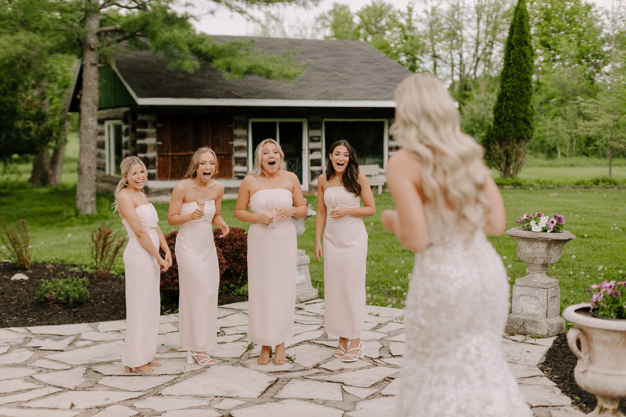 Four women in white dresses celebrate outdoors, with one woman in the foreground facing away. They are holding champagne glasses and standing in front of a rustic house with surrounding greenery.