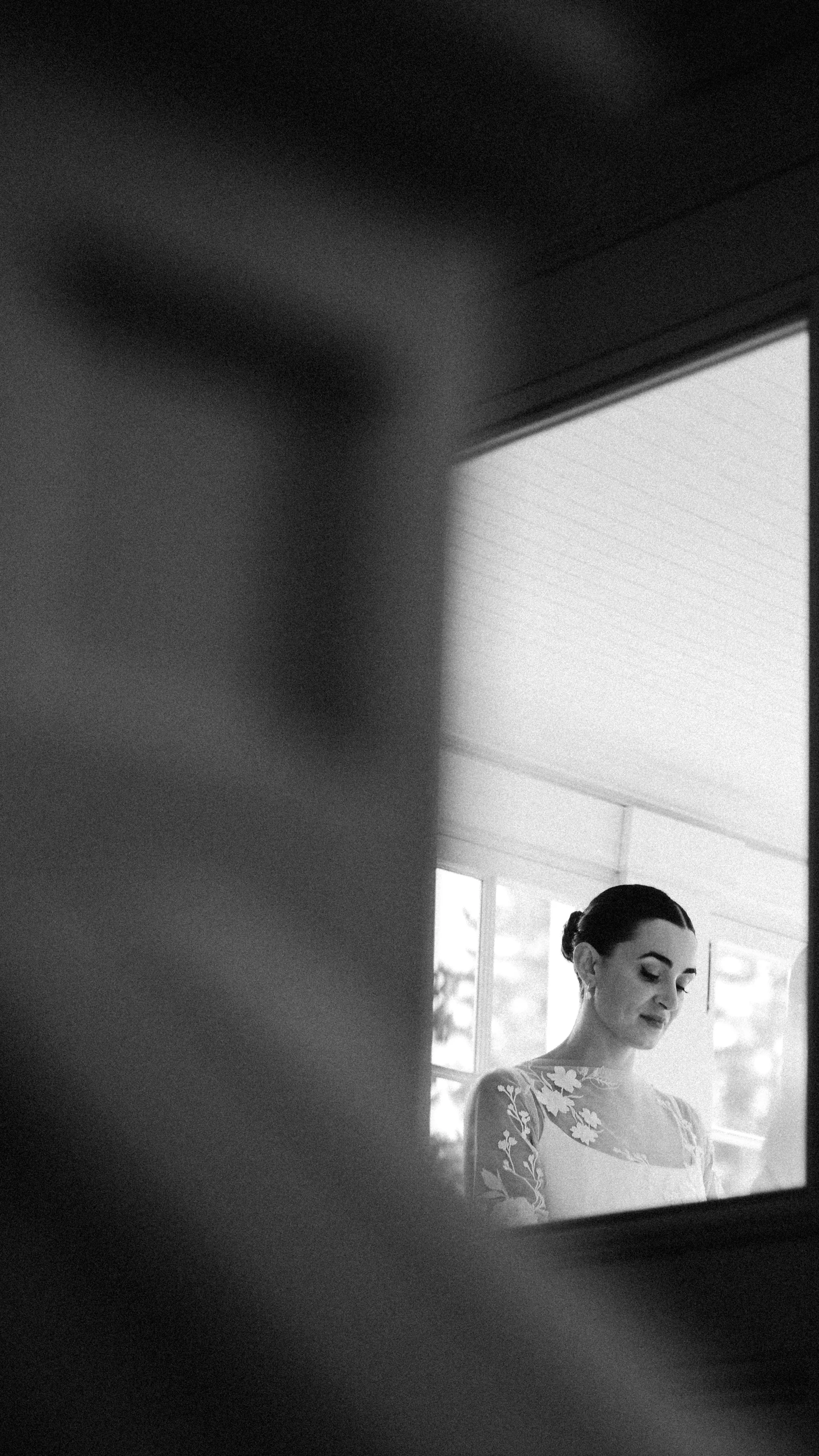 A woman with dark hair styled in an updo, wearing a lace top with floral patterns, standing near a window with blinds, in a black and white photograph.