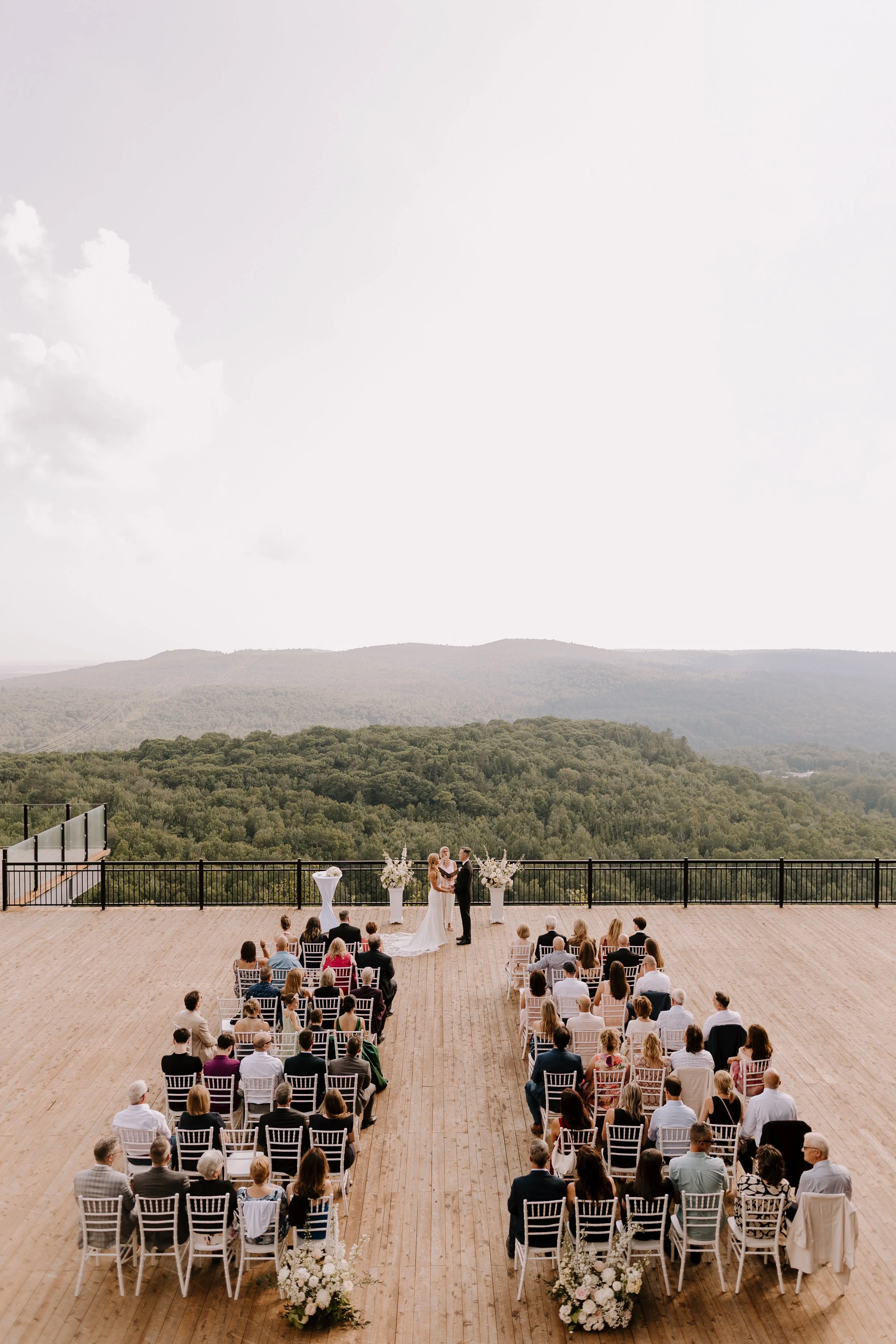 A wedding ceremony on an outdoor wooden deck with a view of green hills and mountains in the background. The bride and groom are standing at the altar, surrounded by officiants and floral arrangements. Guests are seated in rows watching the ceremony.