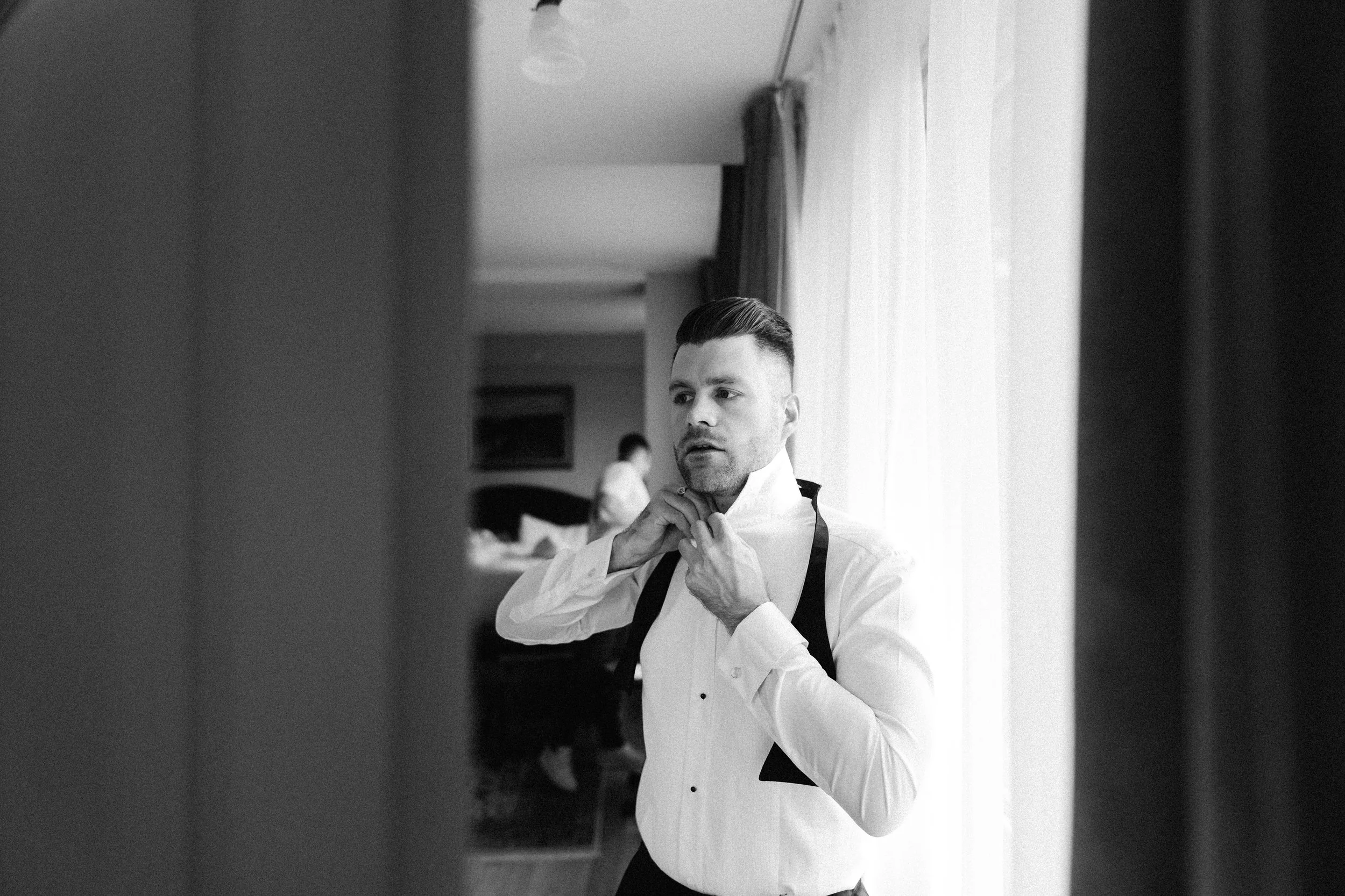A man in a white shirt is adjusting his black tie in front of a mirror, with sunlight coming through a window behind him.