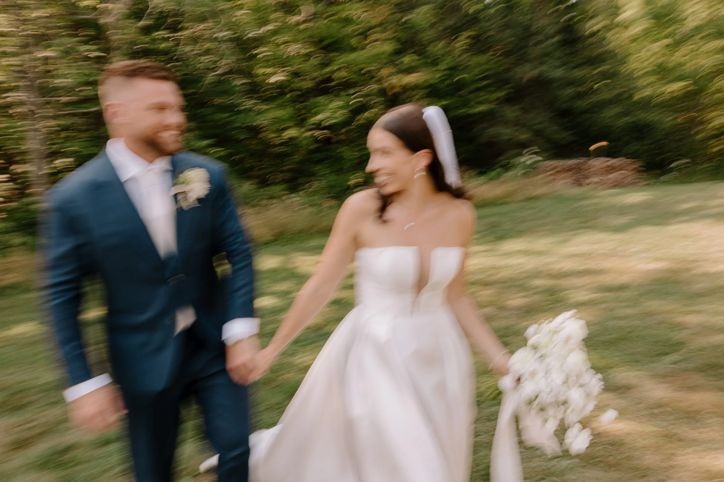 A blurry photo of a man and woman holding hands outdoors, possibly at a wedding, with trees and greenery in the background.