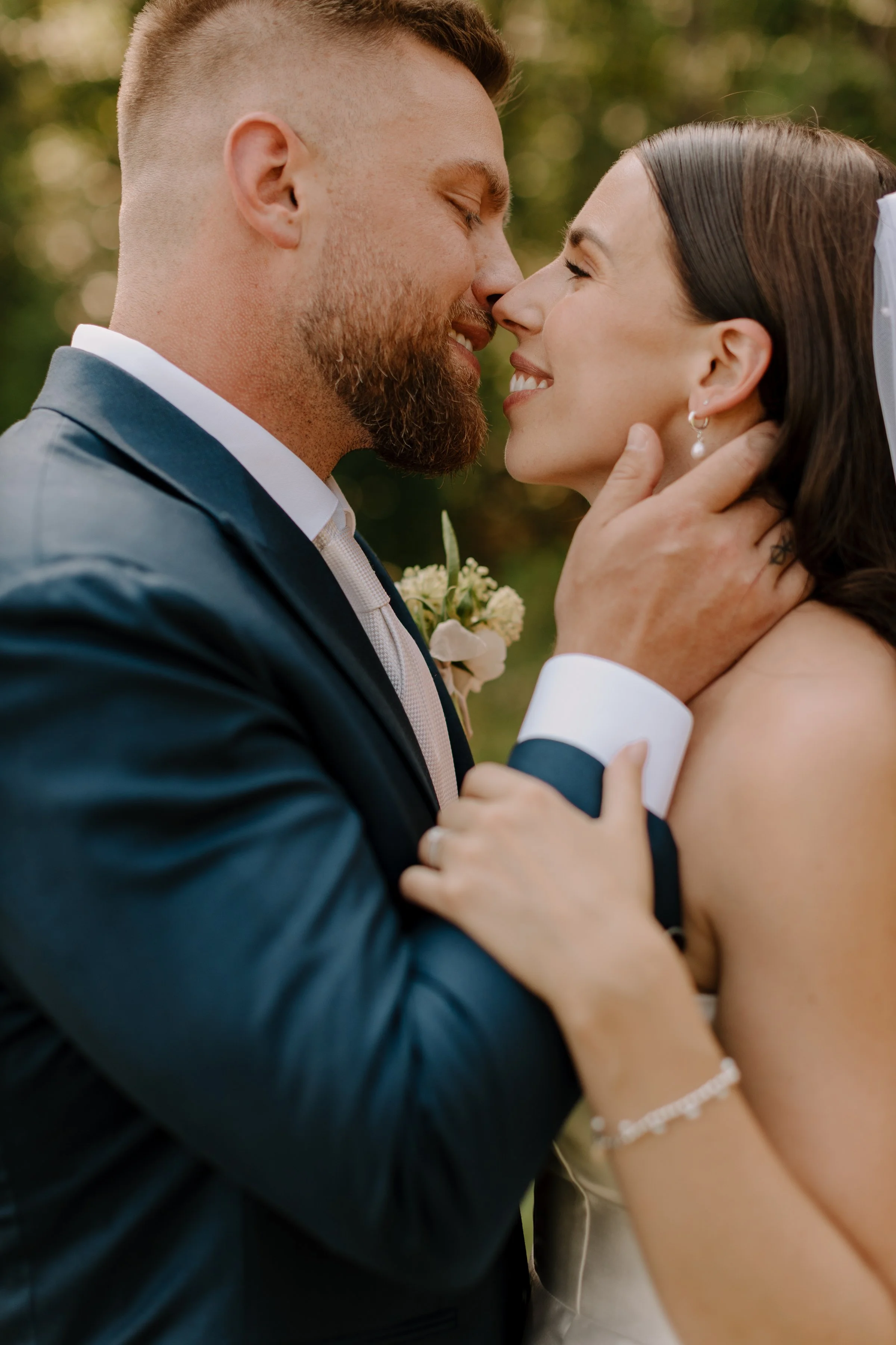 A close-up of a couple, dressed in wedding attire, touching noses and smiling in an outdoor setting during daytime.