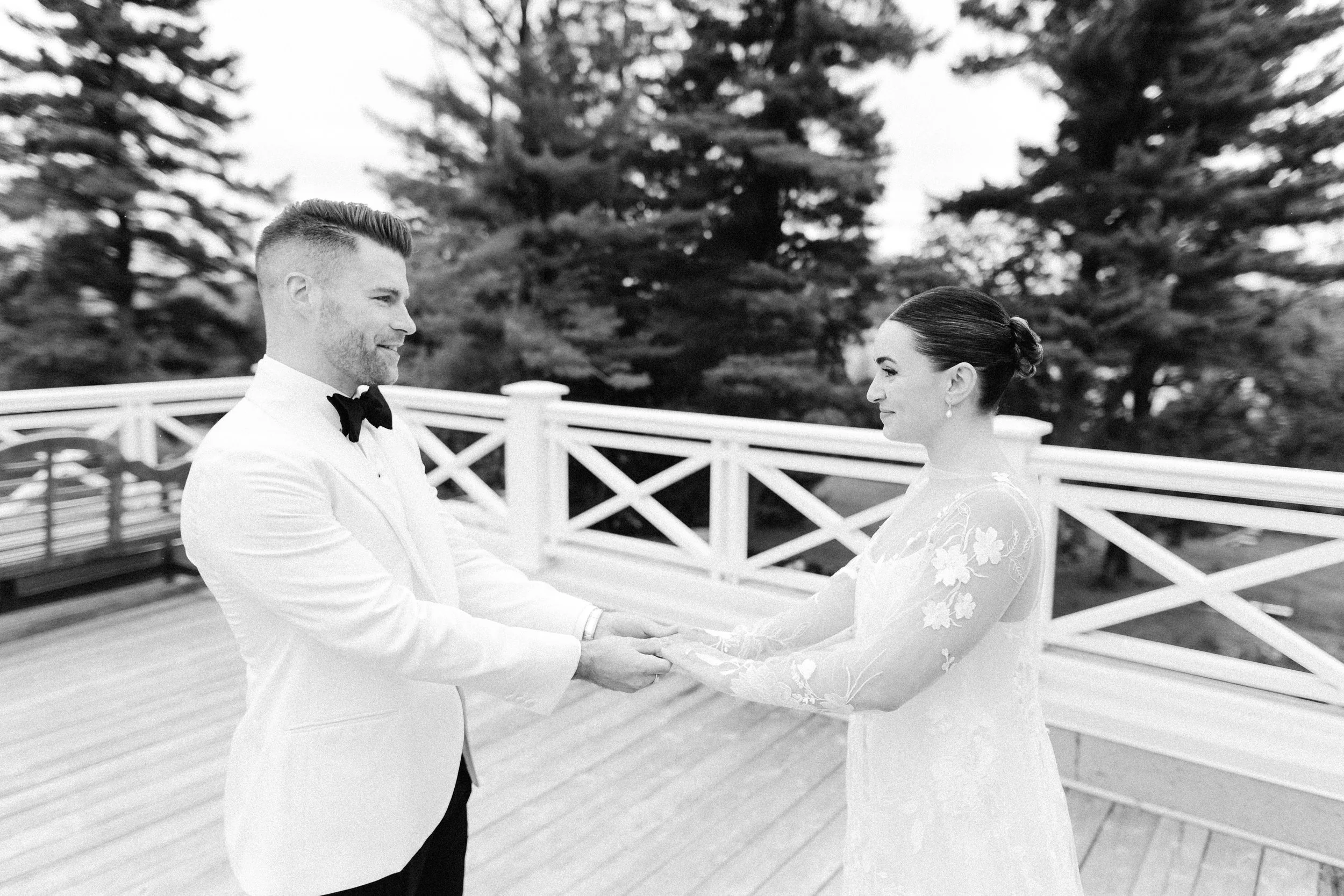 A black and white photo of a bride and groom holding hands and looking at each other outdoors with trees in the background.