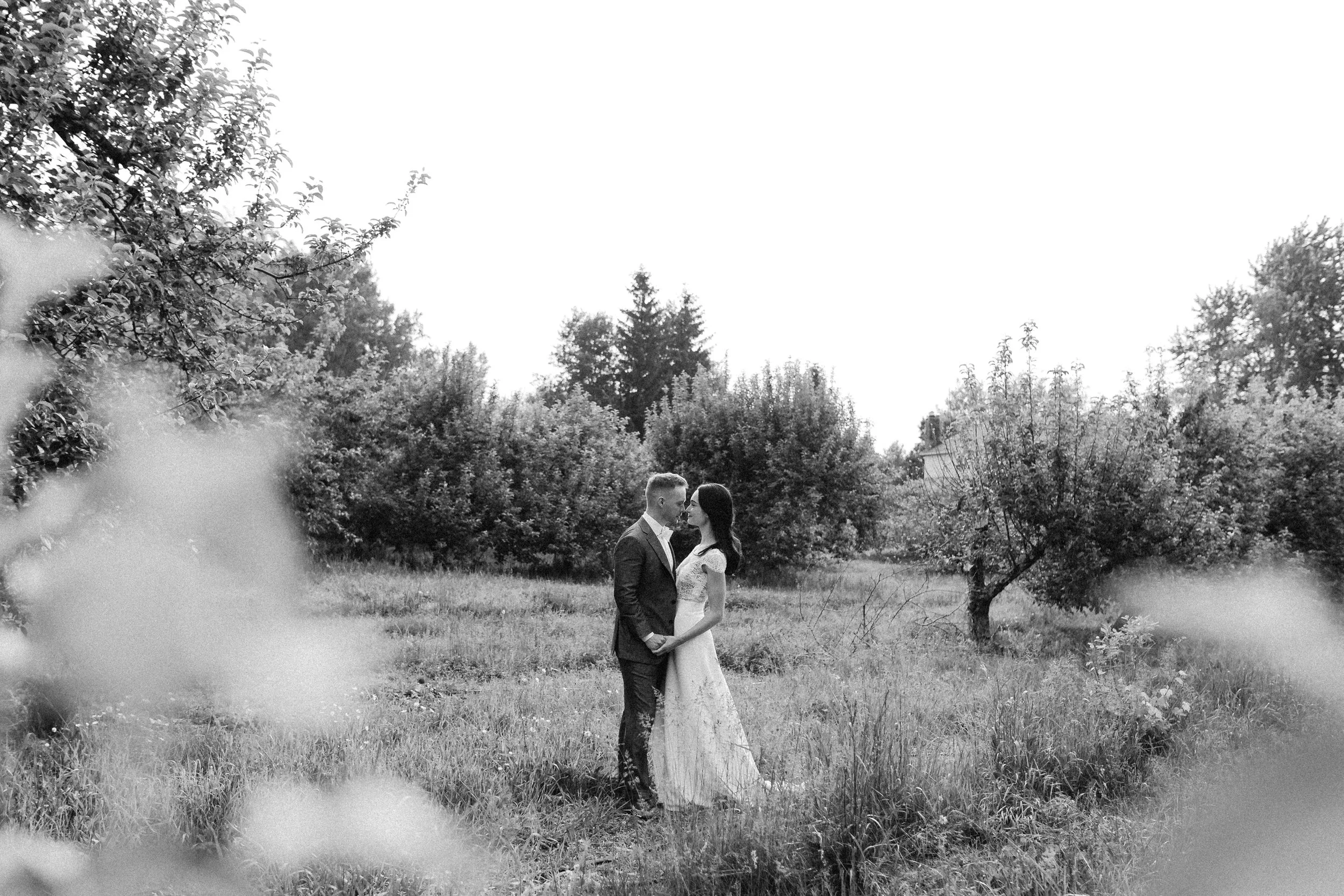 A black-and-white photo of a couple in wedding attire standing close and gazing at each other in a park surrounded by trees and grass.