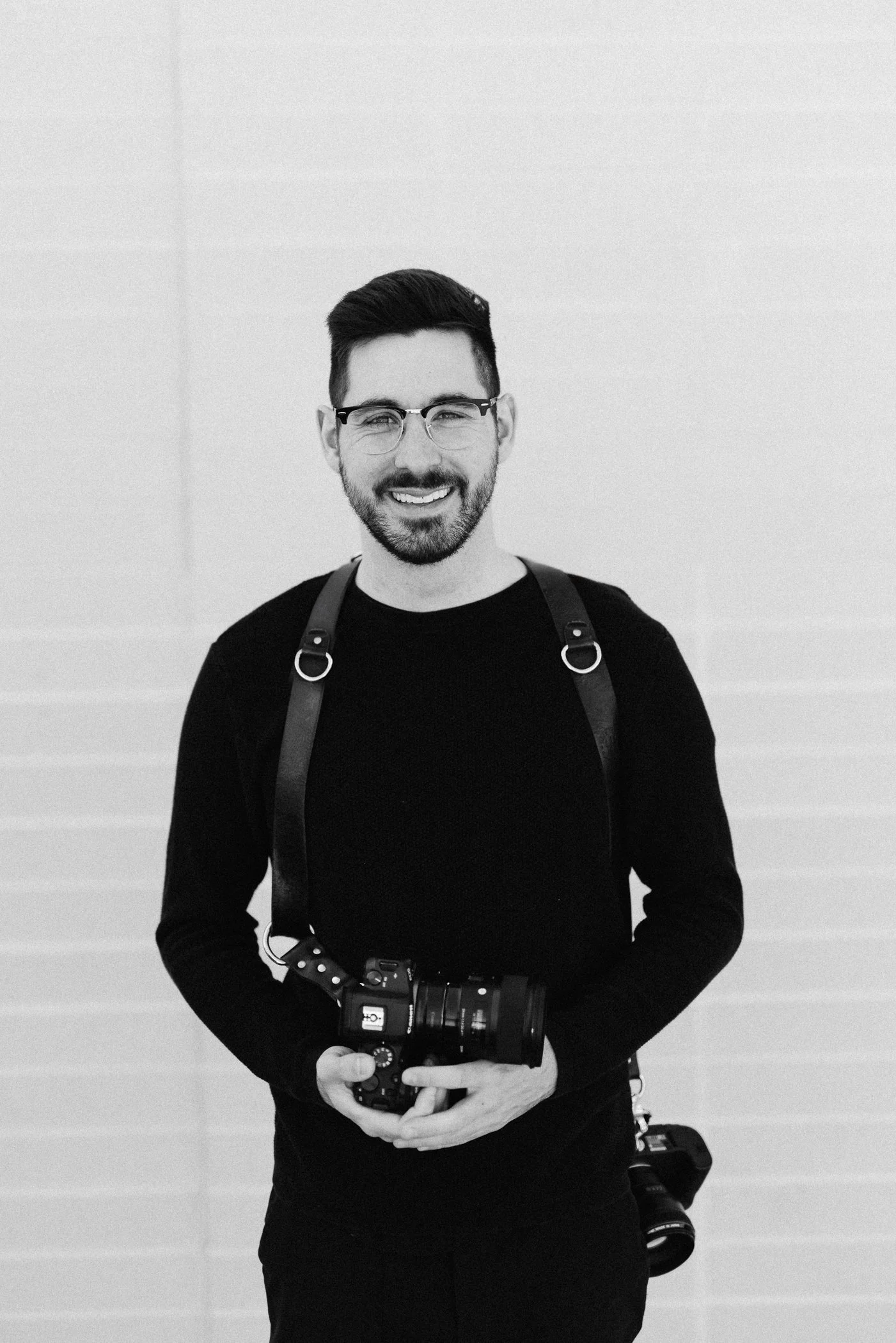 A smiling man with glasses and a beard holding a camera, standing against a plain wall, with another camera hanging from his side.