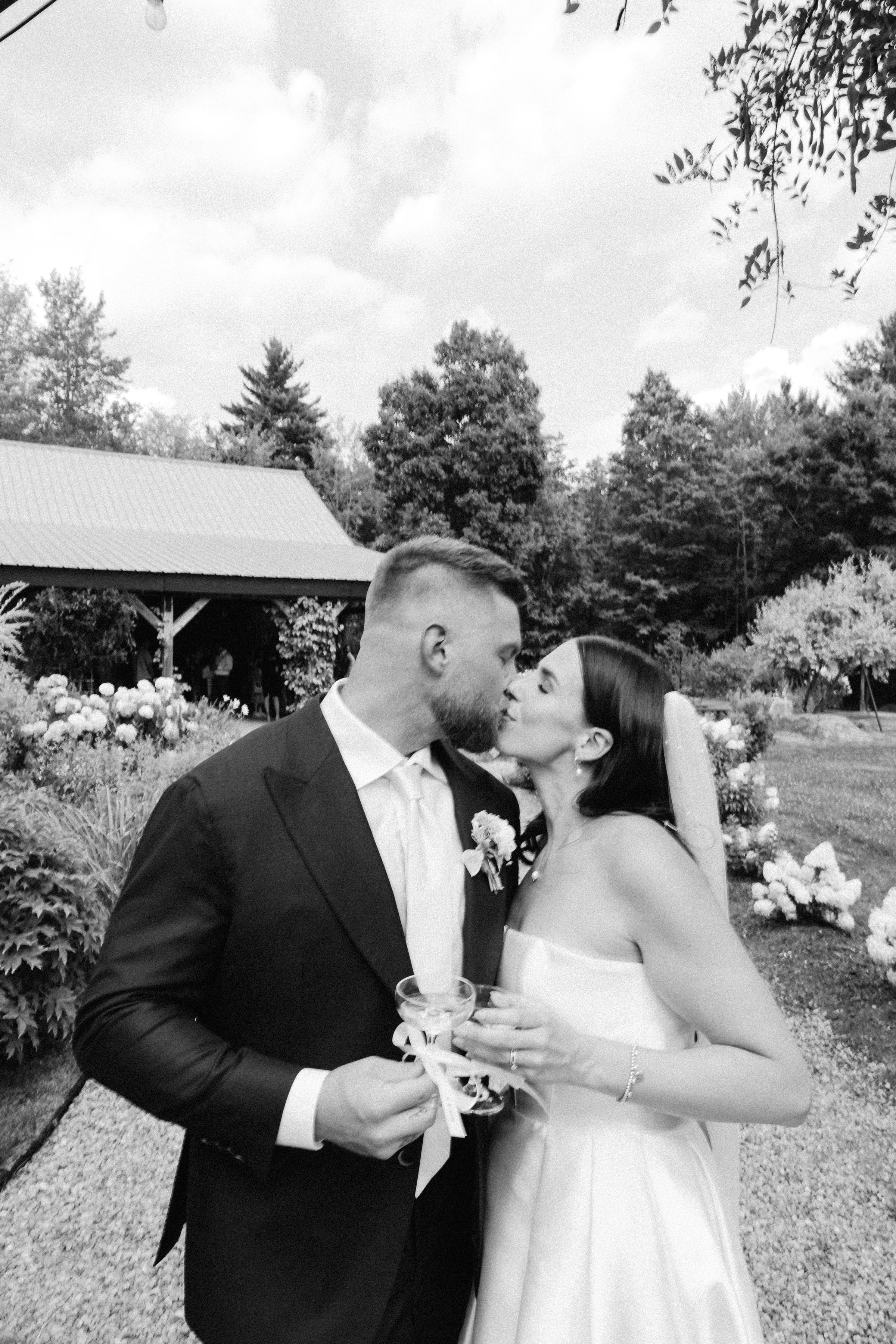 A bride and groom split a kiss during their wedding celebration outdoors, surrounded by trees and flowers.