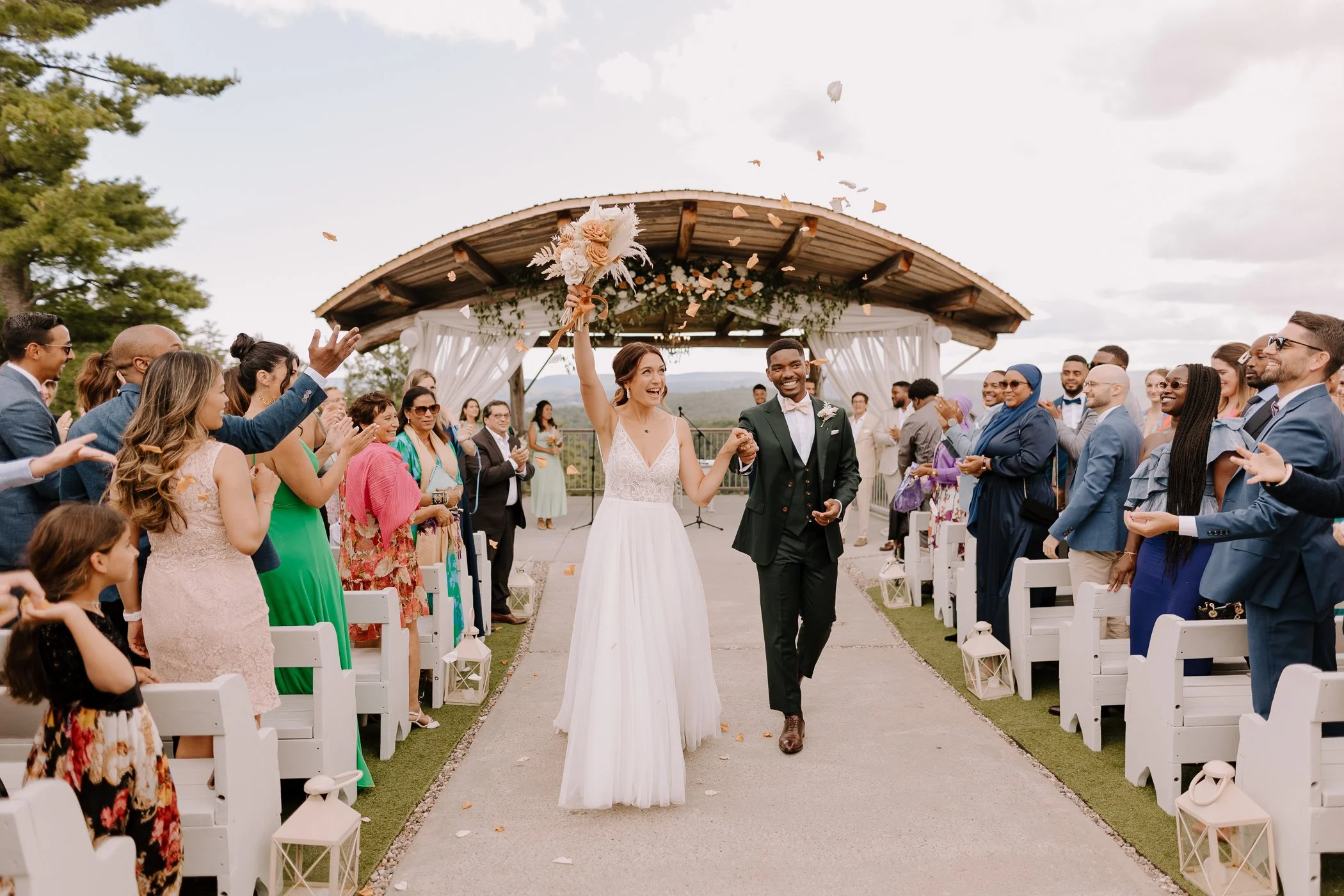 A bride and groom walk down the aisle at an outdoor wedding, guests celebrating and throwing flower petals, with a wooden arch and cloudy sky in the background.
