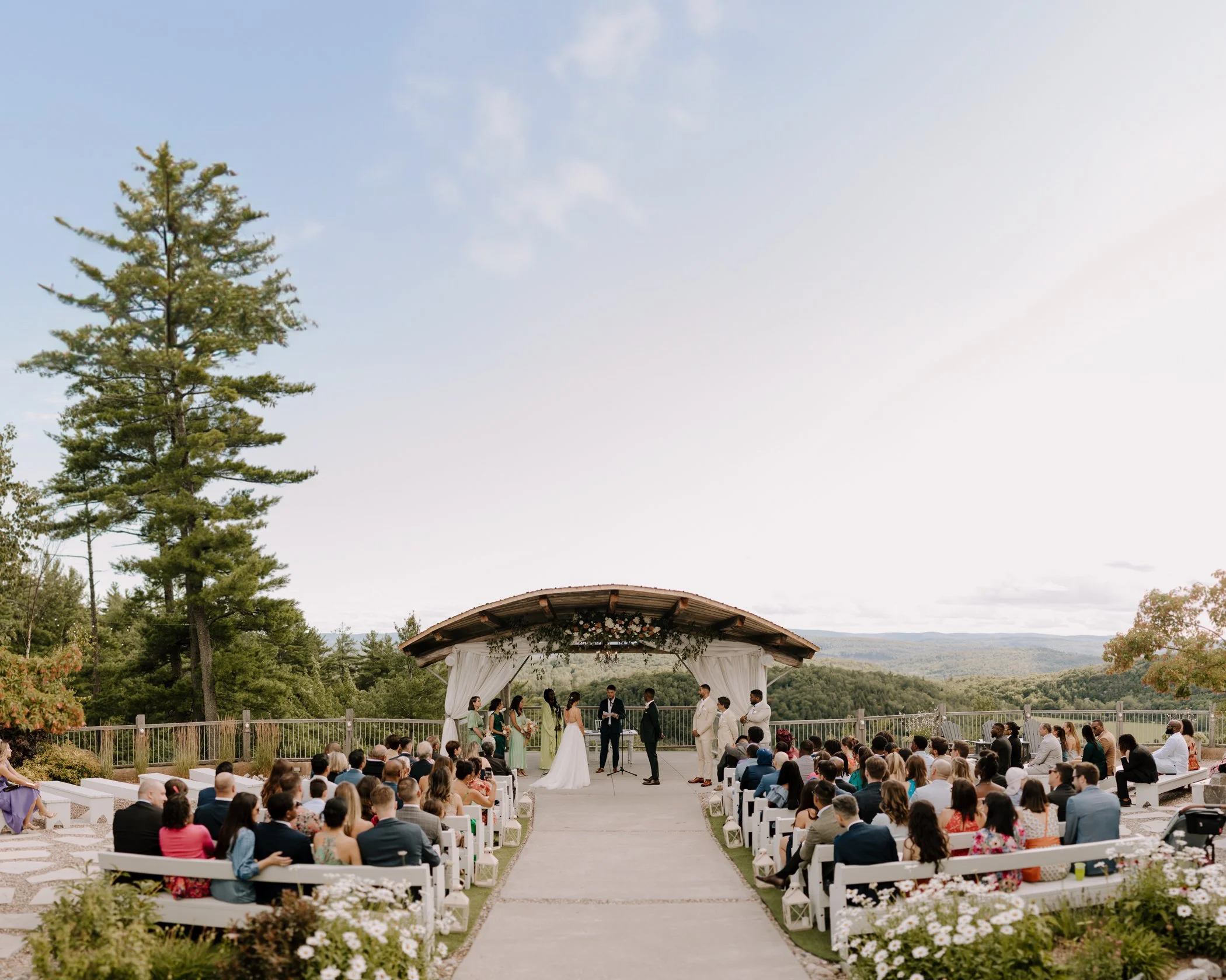 Outdoor wedding ceremony with guests seated watching the bride and groom exchange vows under a canopy with floral decorations, set against a scenic natural landscape.