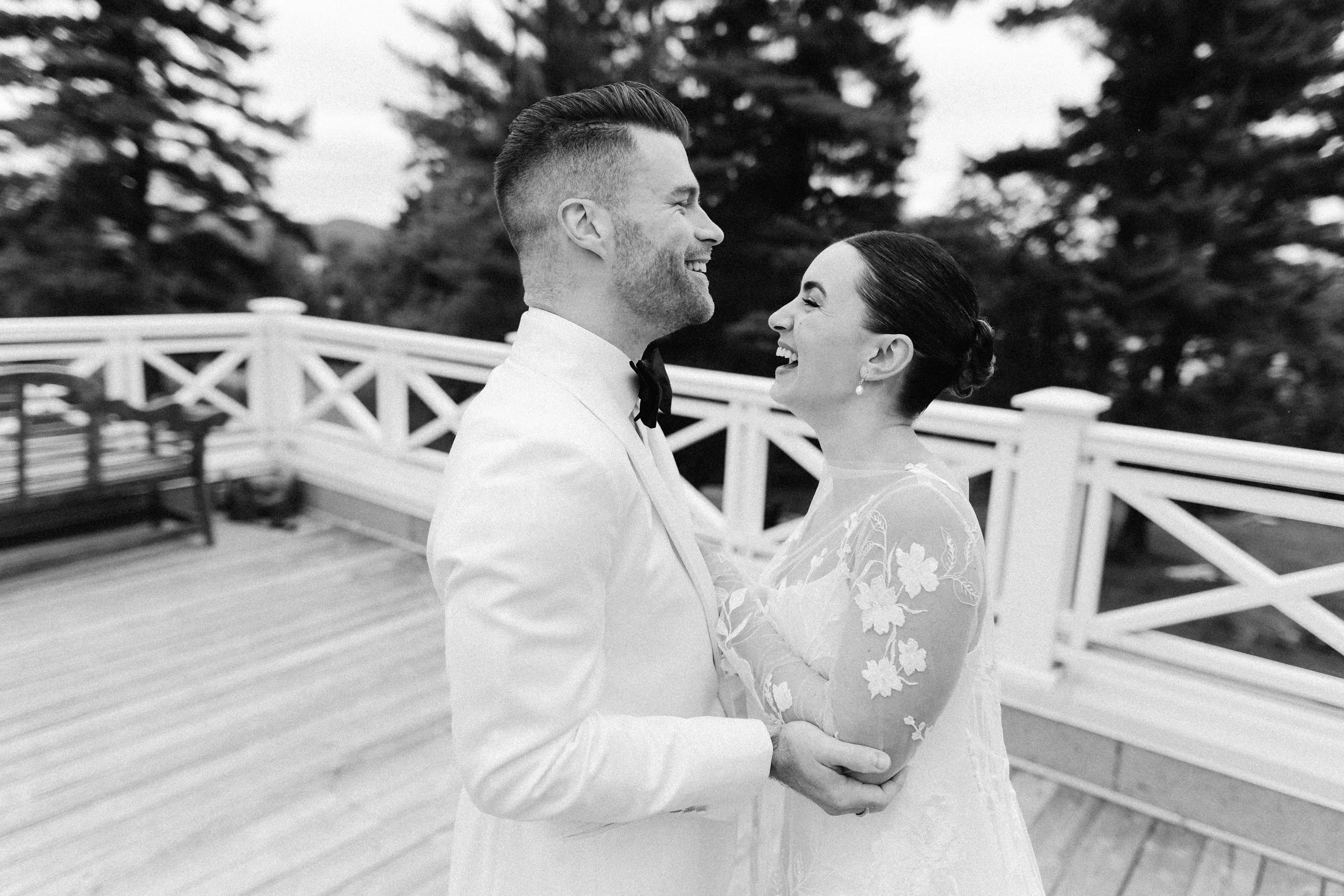 A black-and-white photo of a smiling couple on their wedding day, standing on a wooden deck with a white fence and trees in the background.