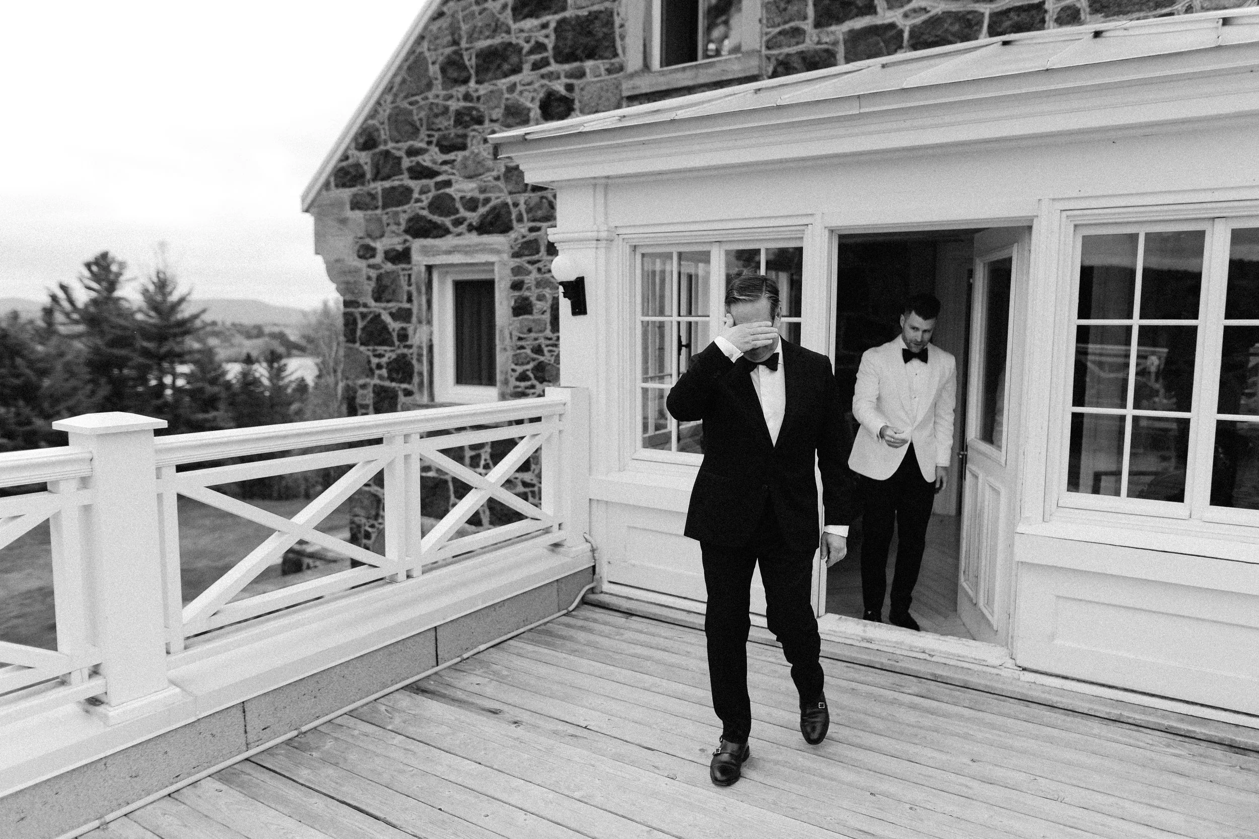 Black and white photo of two men dressed in tuxedos walking out of a house onto a wooden porch. One man is covering his face with his hand while the other man smiles.