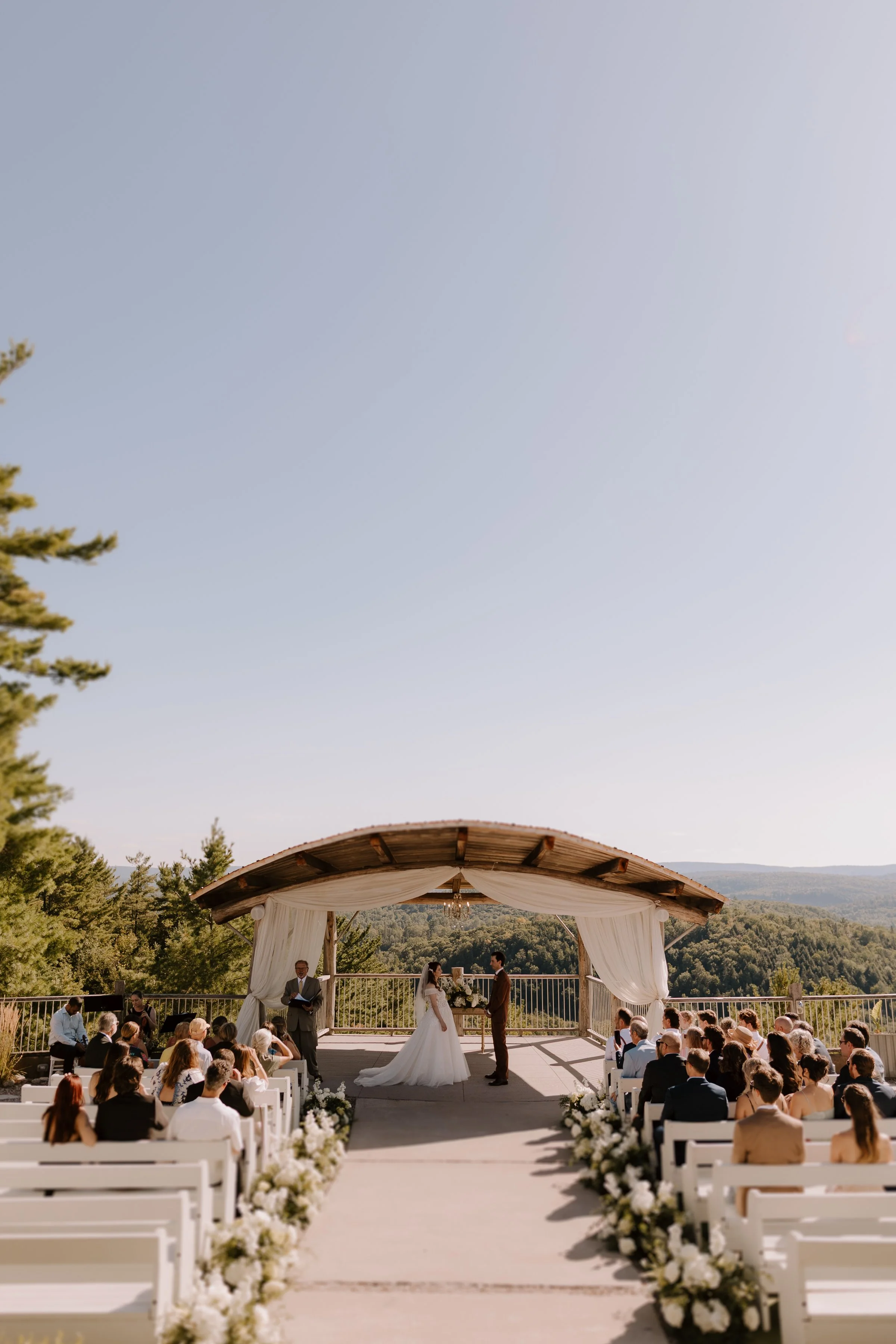Outdoor wedding ceremony under a wooden pavilion with white drapery, floral arrangements, and guests seated on white chairs, against a backdrop of mountains and trees.