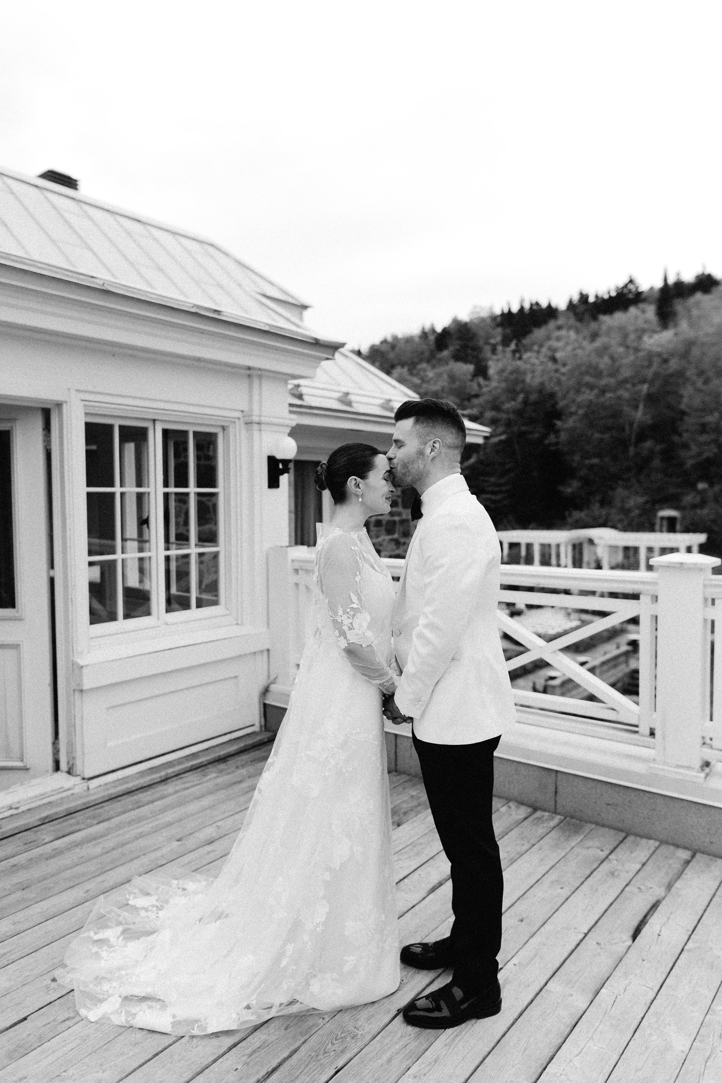 A bride and groom standing close together on a wooden deck, sharing a kiss outdoors during their wedding. The bride wears a lace wedding dress with long sleeves, and the groom is dressed in a tuxedo with a bowtie. There are trees and a white house wi