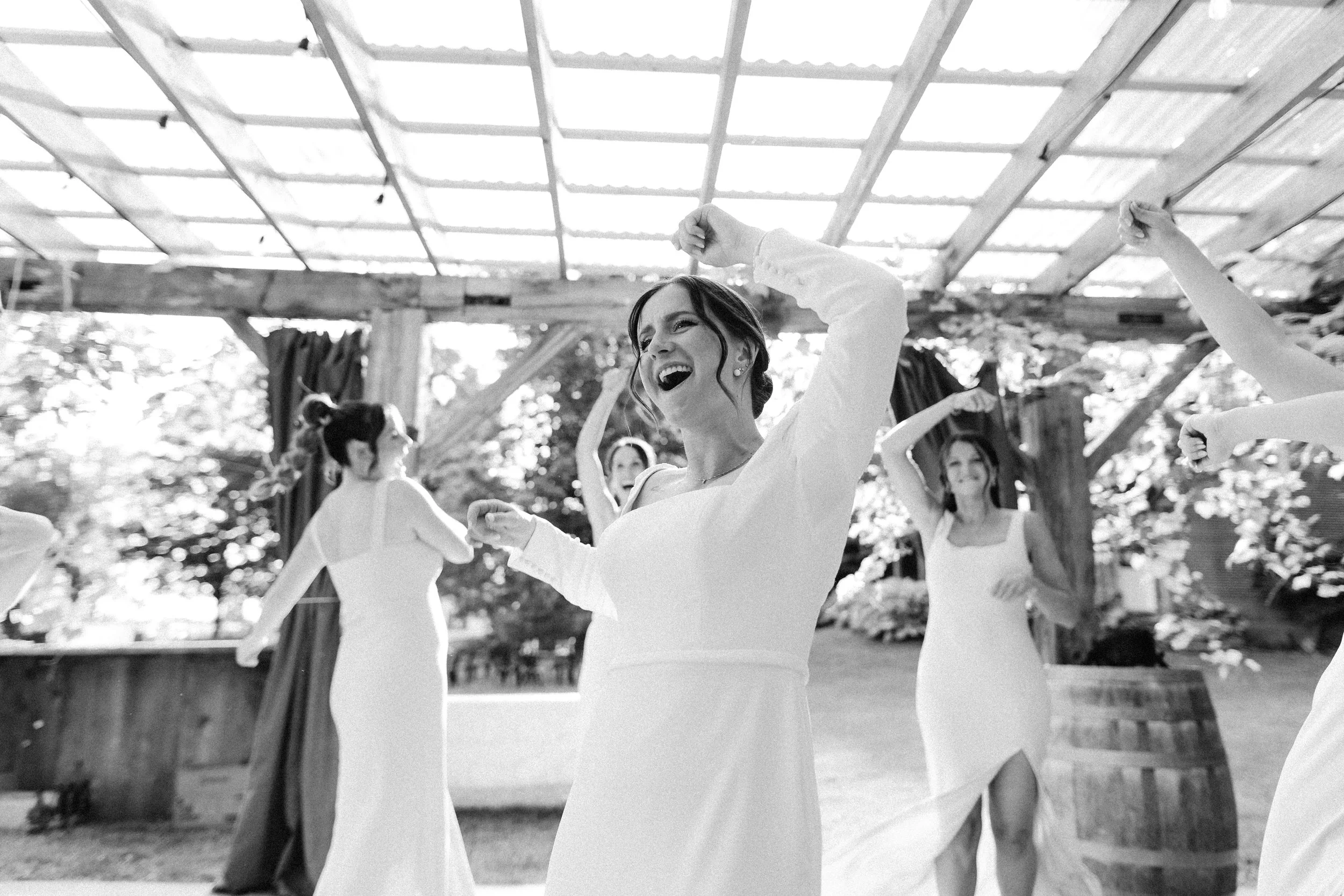 Women in flowy dresses dancing outdoors during daylight under a wooden pergola