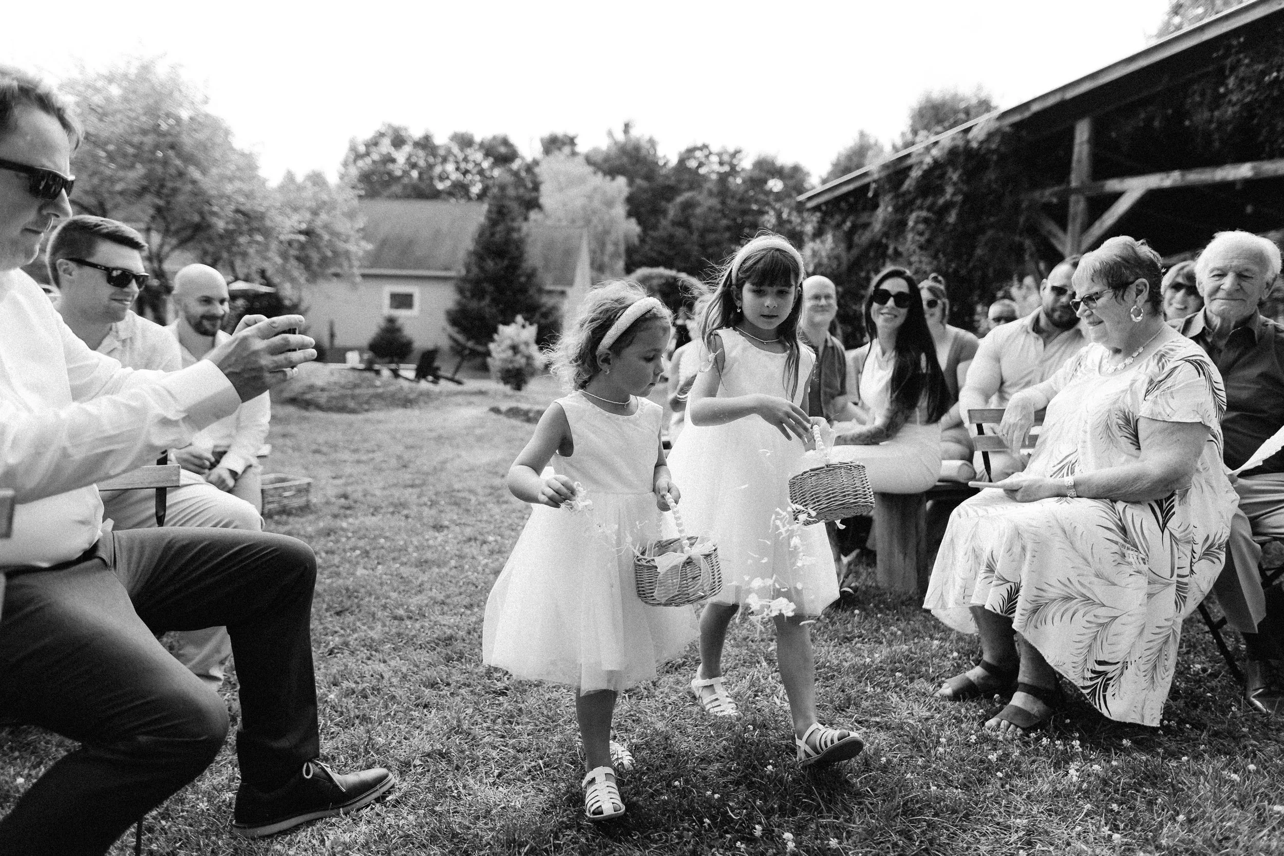 A black and white photo of a group gathering outdoors, with two young girls holding baskets, surrounded by seated adults.