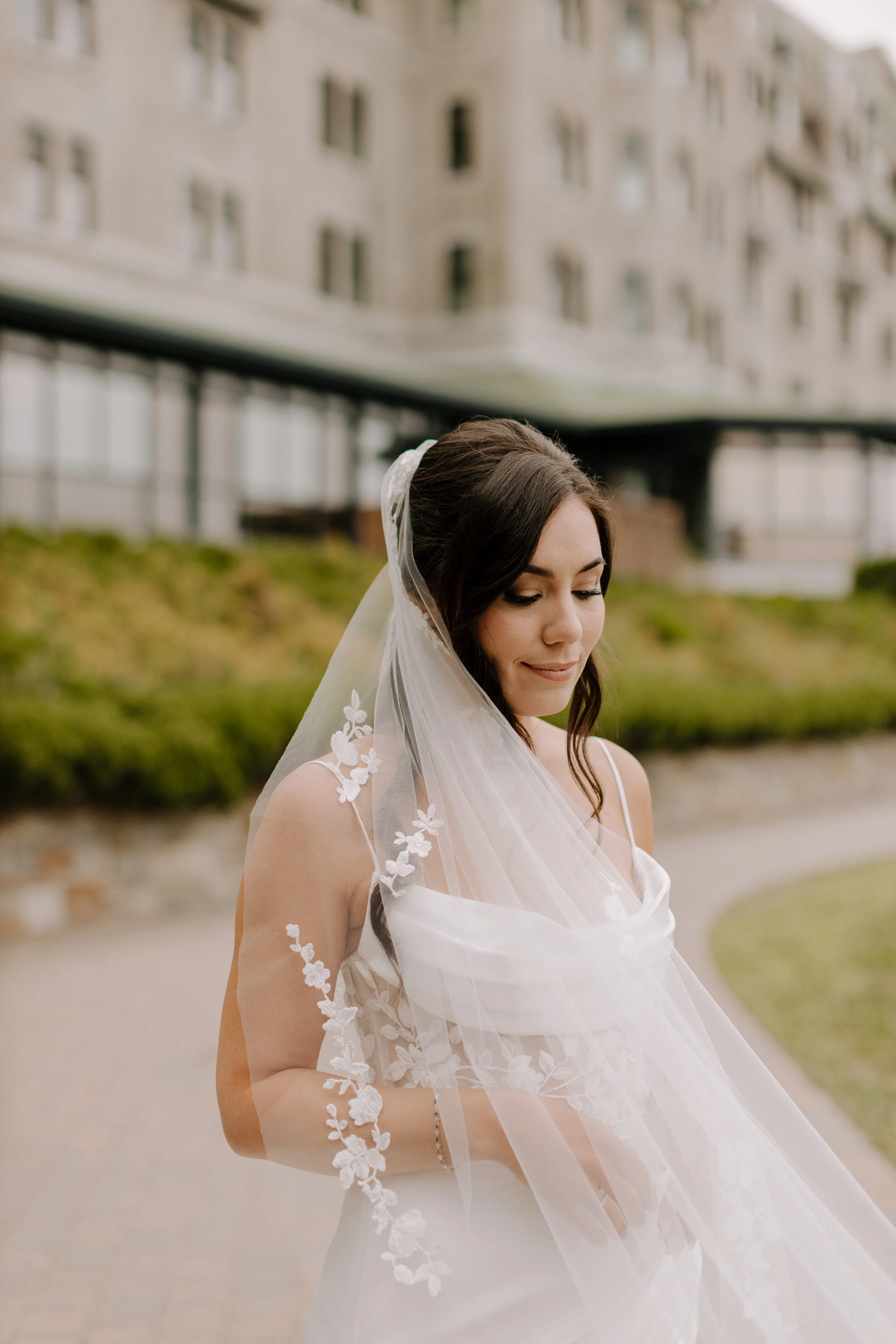 A bride standing outdoors near a building with multiple windows, wearing a white wedding dress and veil with floral embroidery, looking down with a gentle smile.