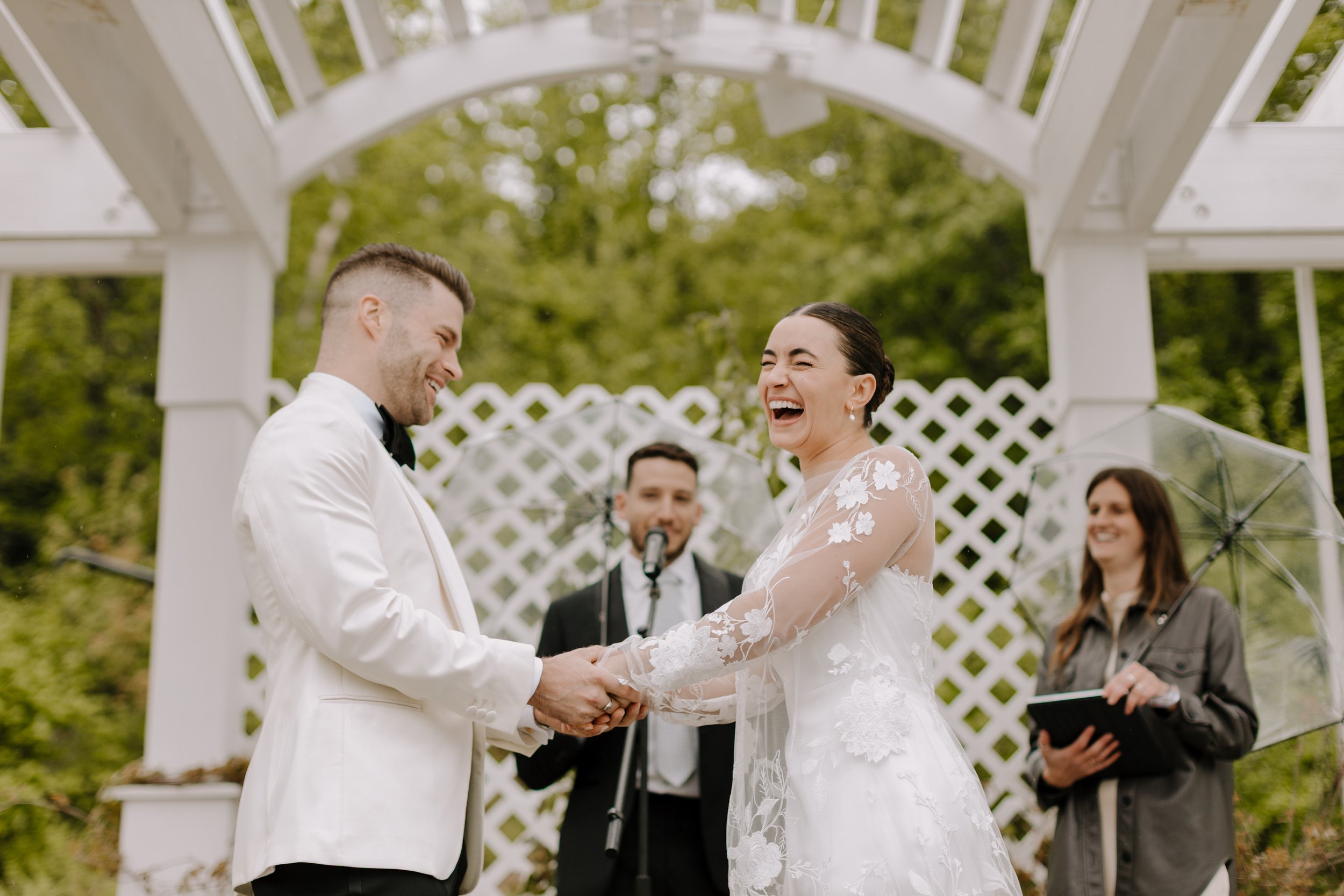 A couple gets married outdoors, holding hands and smiling at each other, with a officiant and guests in the background under a white garden arch.