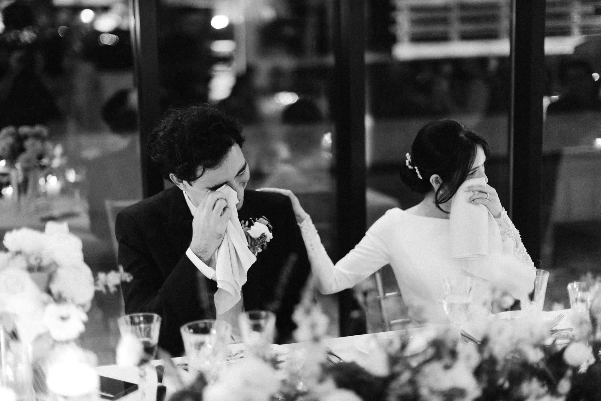 Black and white photo of a bride and groom sitting at a table, wiping tears during a wedding celebration.