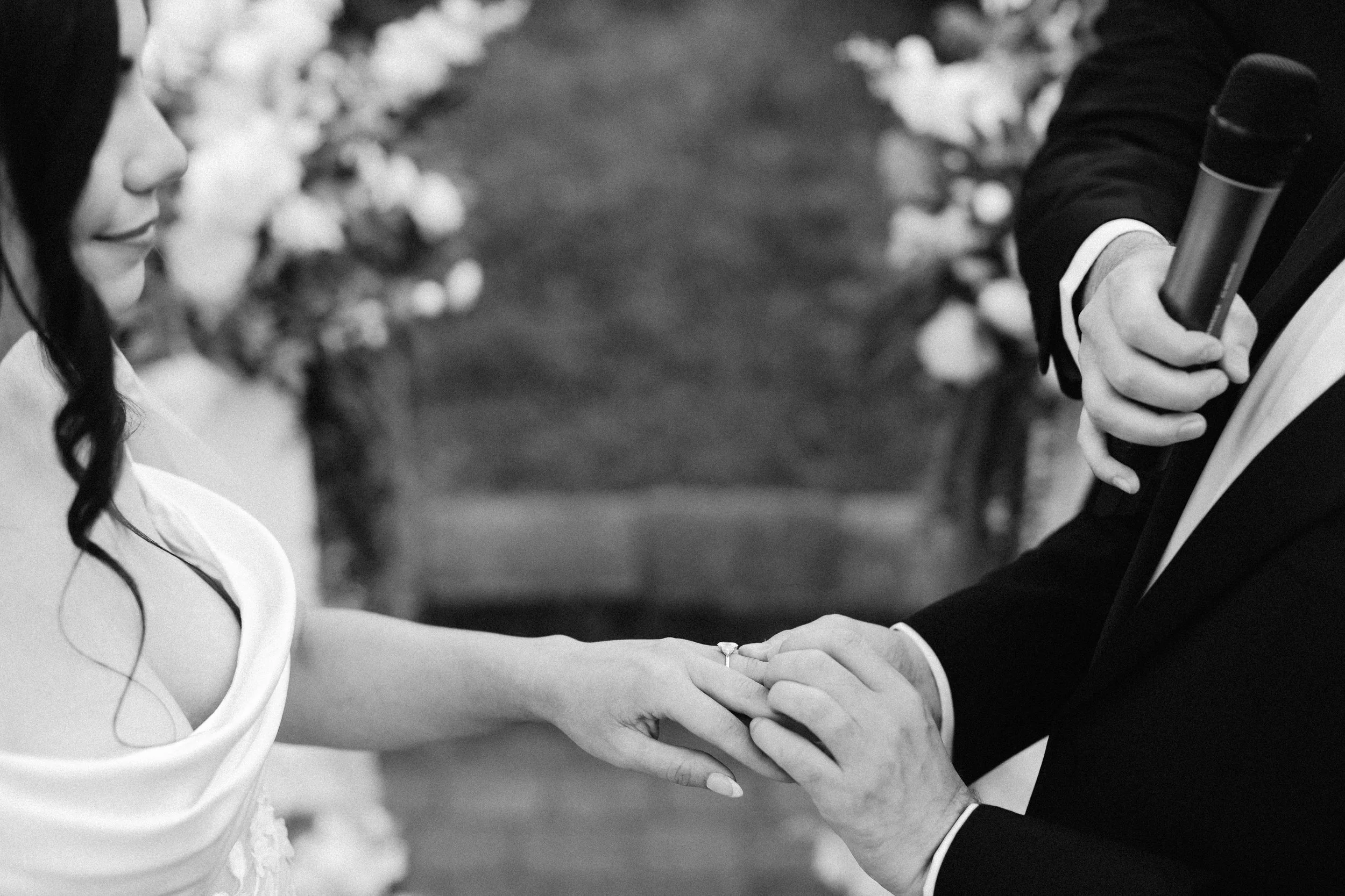 A black and white photograph showing a woman in a white dress and a man in a dark suit holding hands, with the woman's face partially visible, looking at the man and smiling. The background is blurred with bokeh lighting.