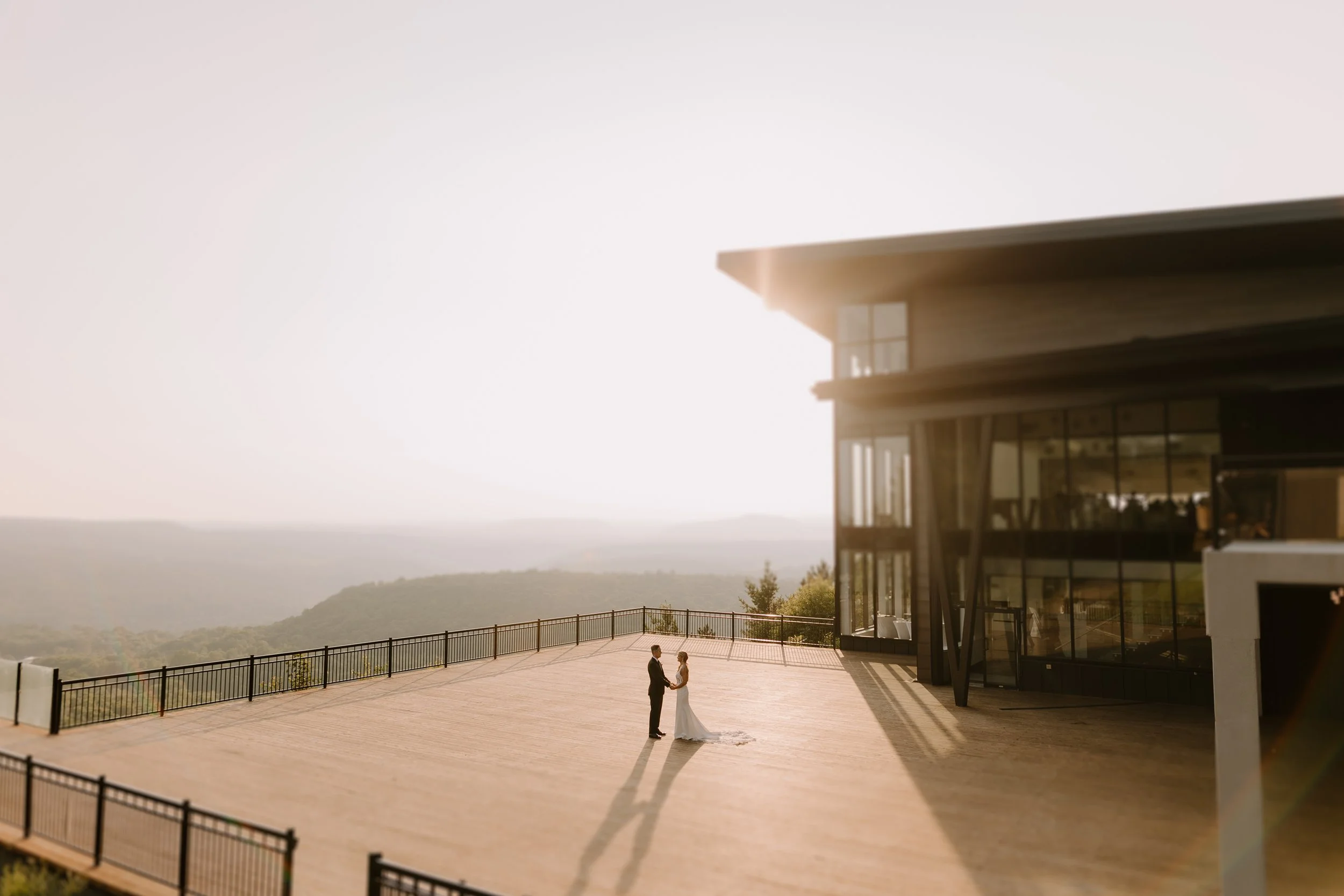 A bride and groom holding hands and looking at each other on a large outdoor terrace with scenic hills in the background, near a modern building with large windows, during sunset.