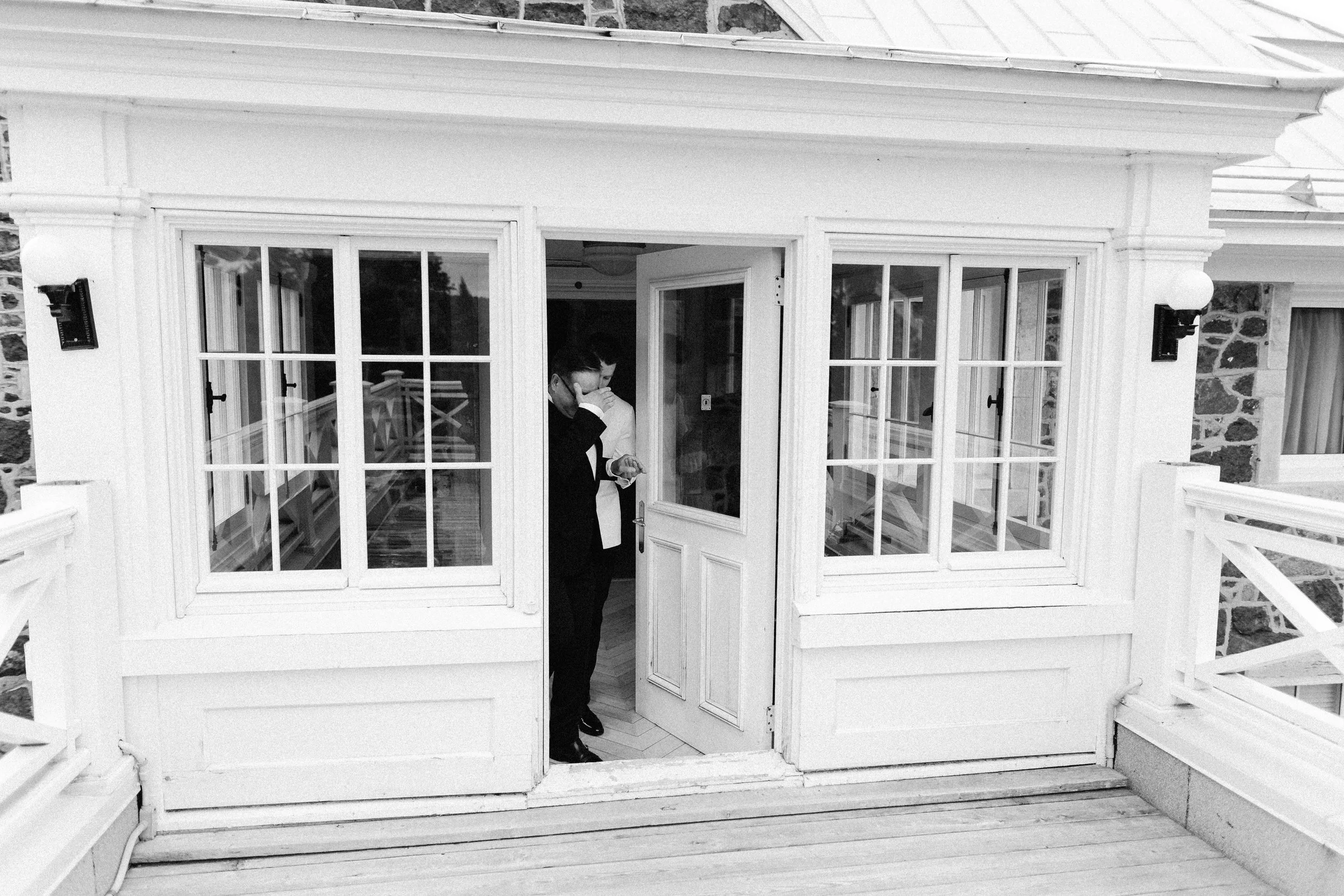 A man in a suit standing inside a glass enclosed porch or sunroom, looking at his phone, with his hand partially covering his face.