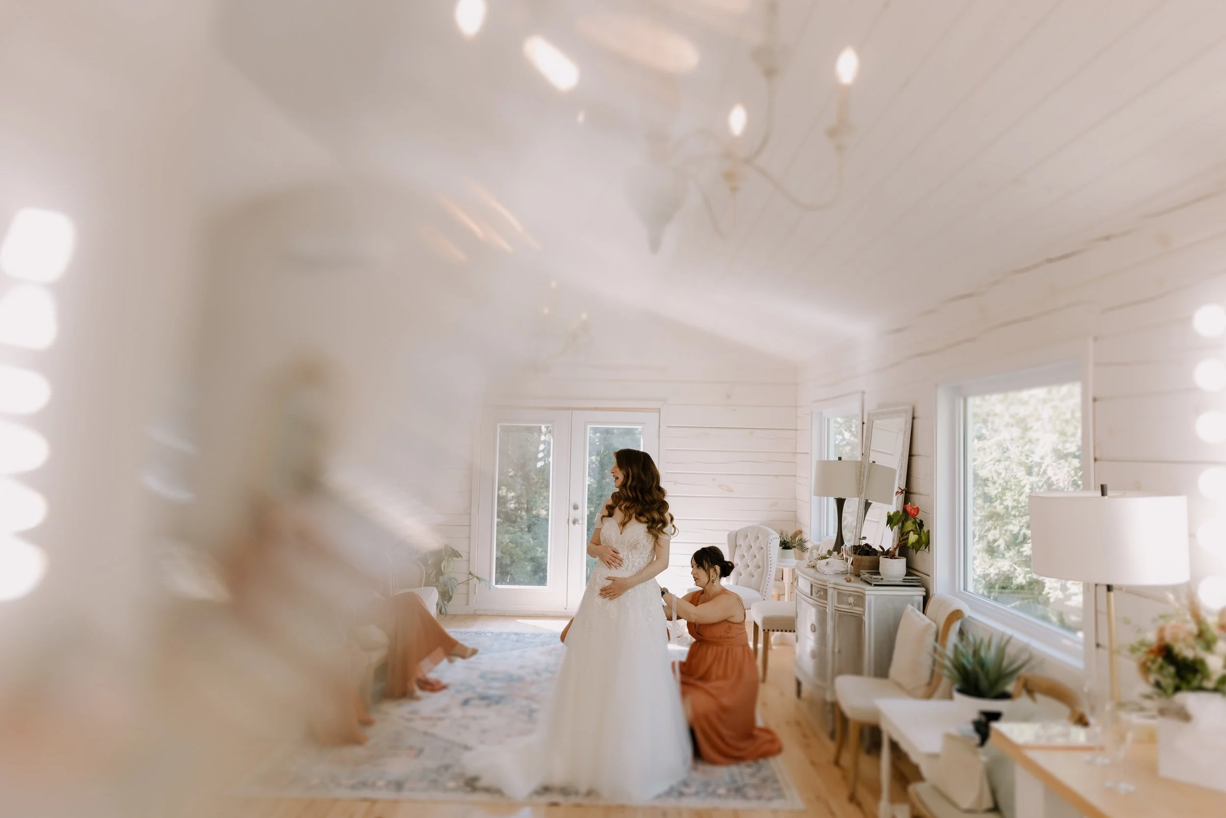 A bride in a white wedding dress getting ready inside a bright, white-walled room with large windows, surrounded by friends, with decorative furniture and plants.