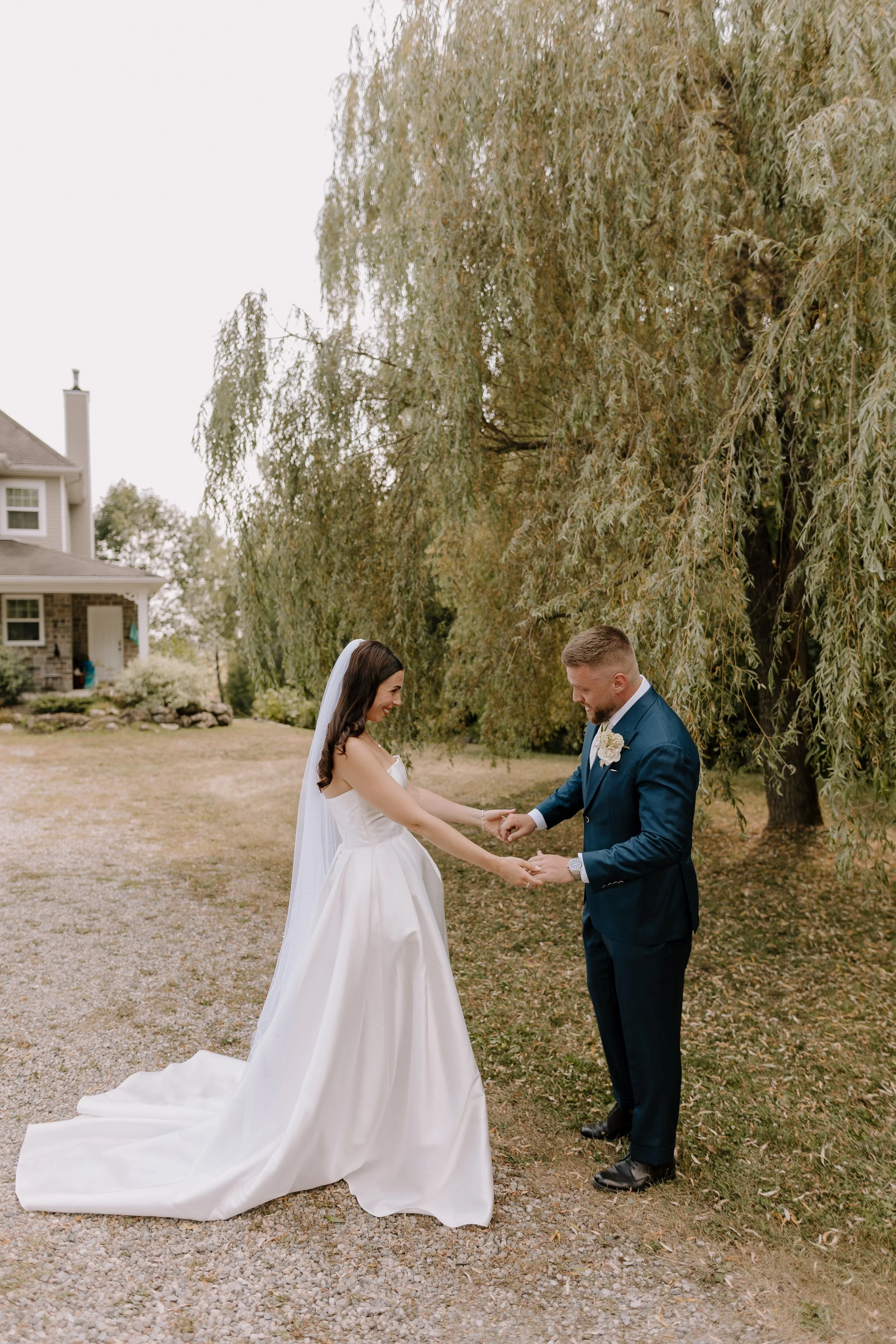 A bride and groom holding hands and smiling during their wedding ceremony outdoors, with a large tree and a house in the background.