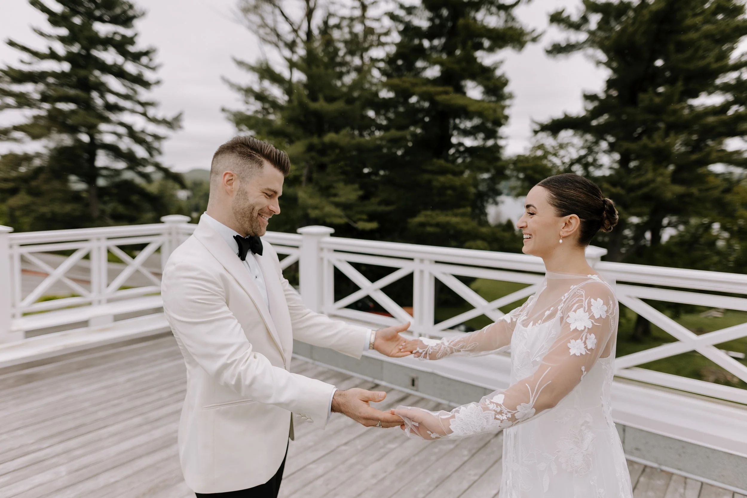 A bride and groom holding hands and smiling during their outdoor wedding ceremony on a cloudy day, with tall trees in the background and a white wooden railing.