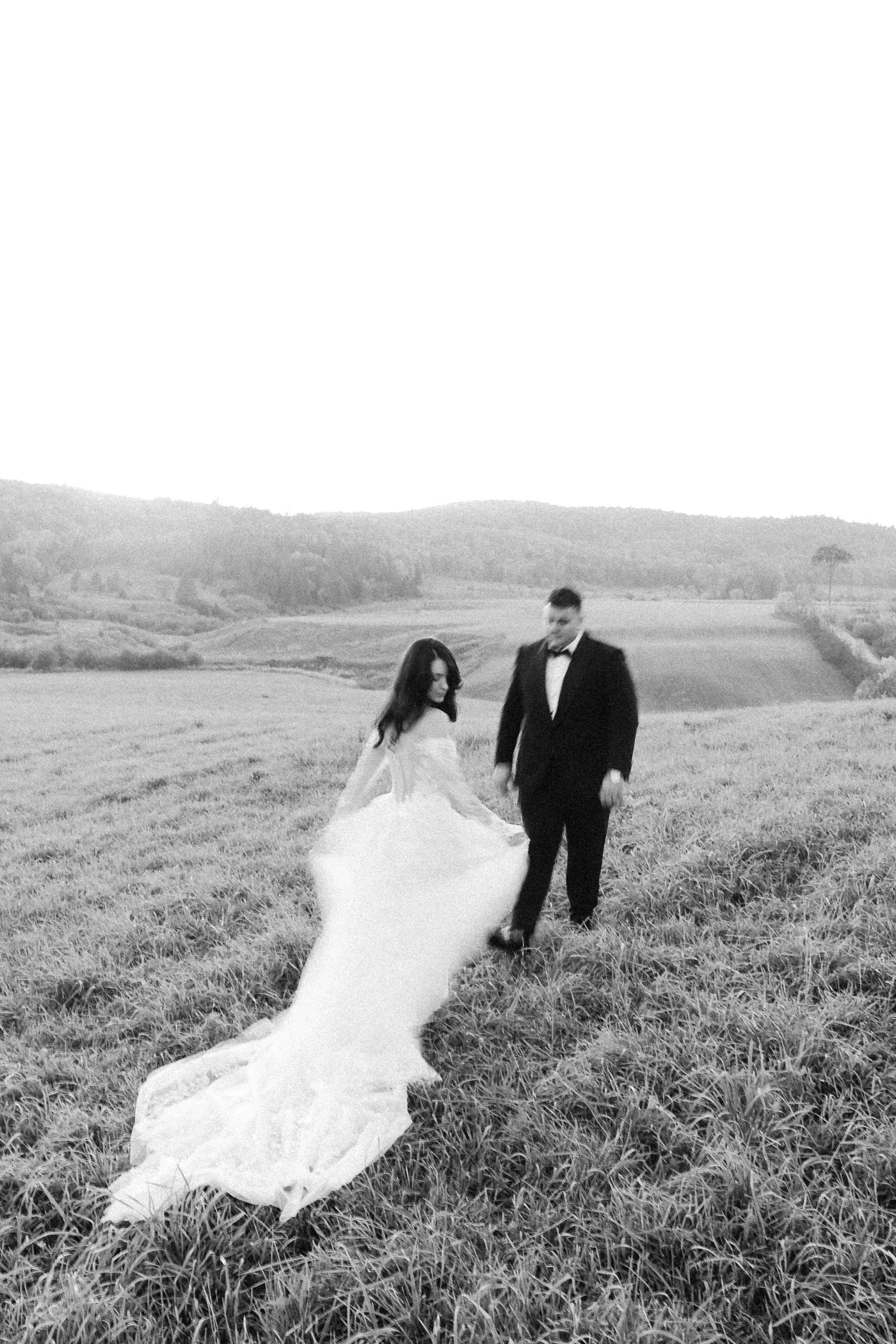 A bride and groom walk hand in hand across a grassy field with rolling hills in the background, the bride’s long veil trailing behind her.