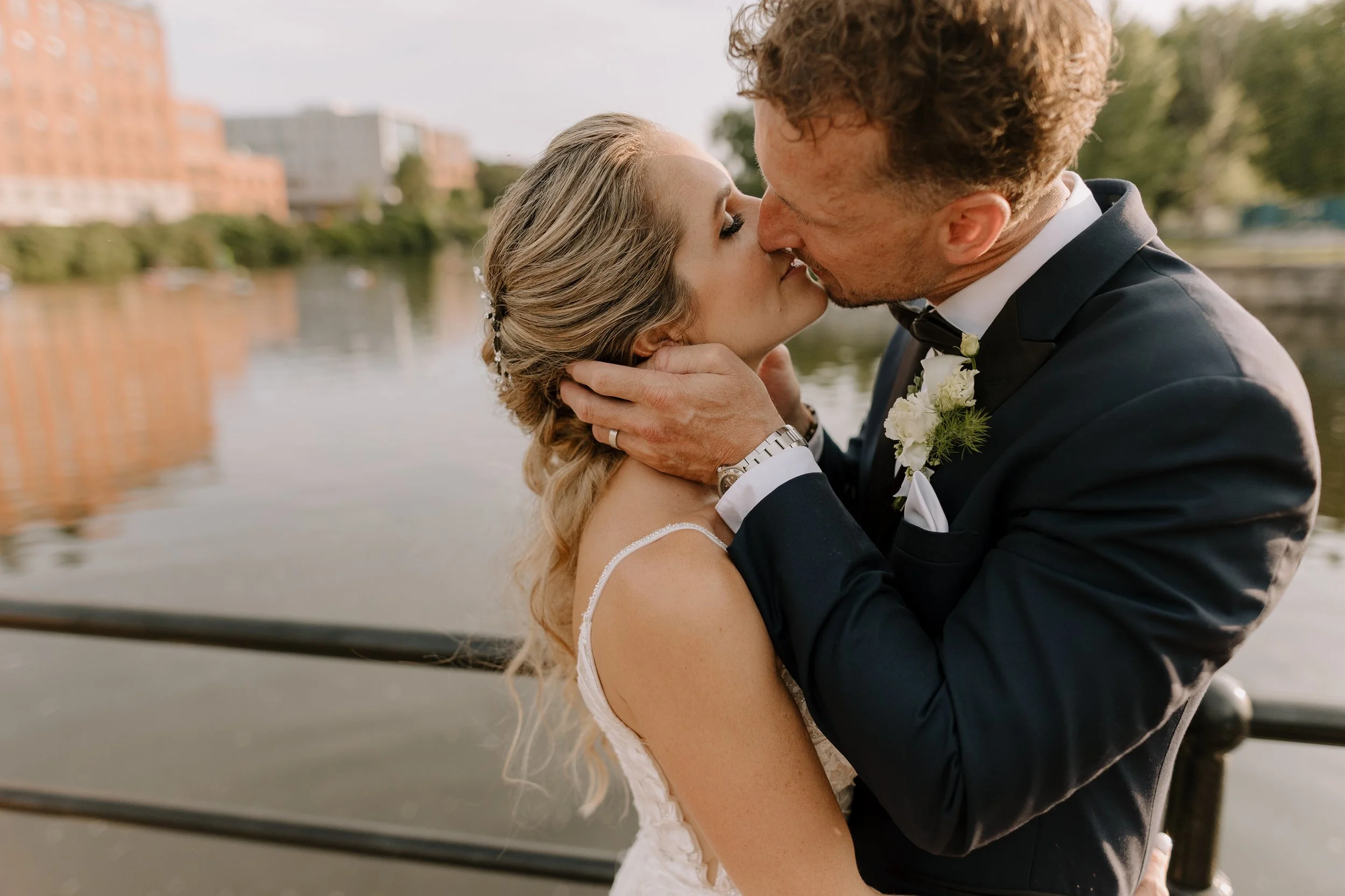 A bride and groom kissing outdoors near water with buildings in the background.