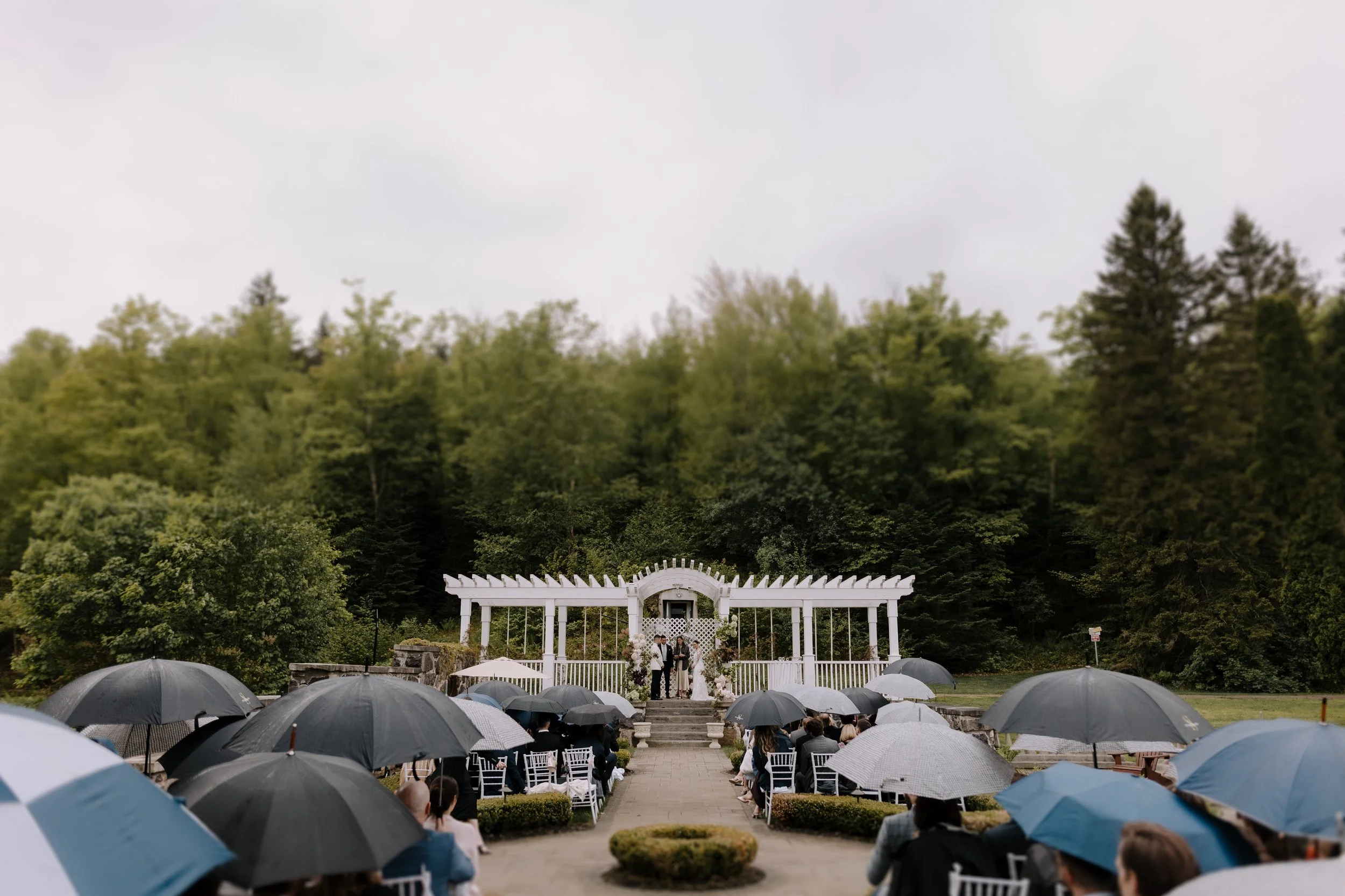 Outdoor wedding ceremony with guests holding umbrellas, a white archway, and a wooded backdrop on a cloudy day.