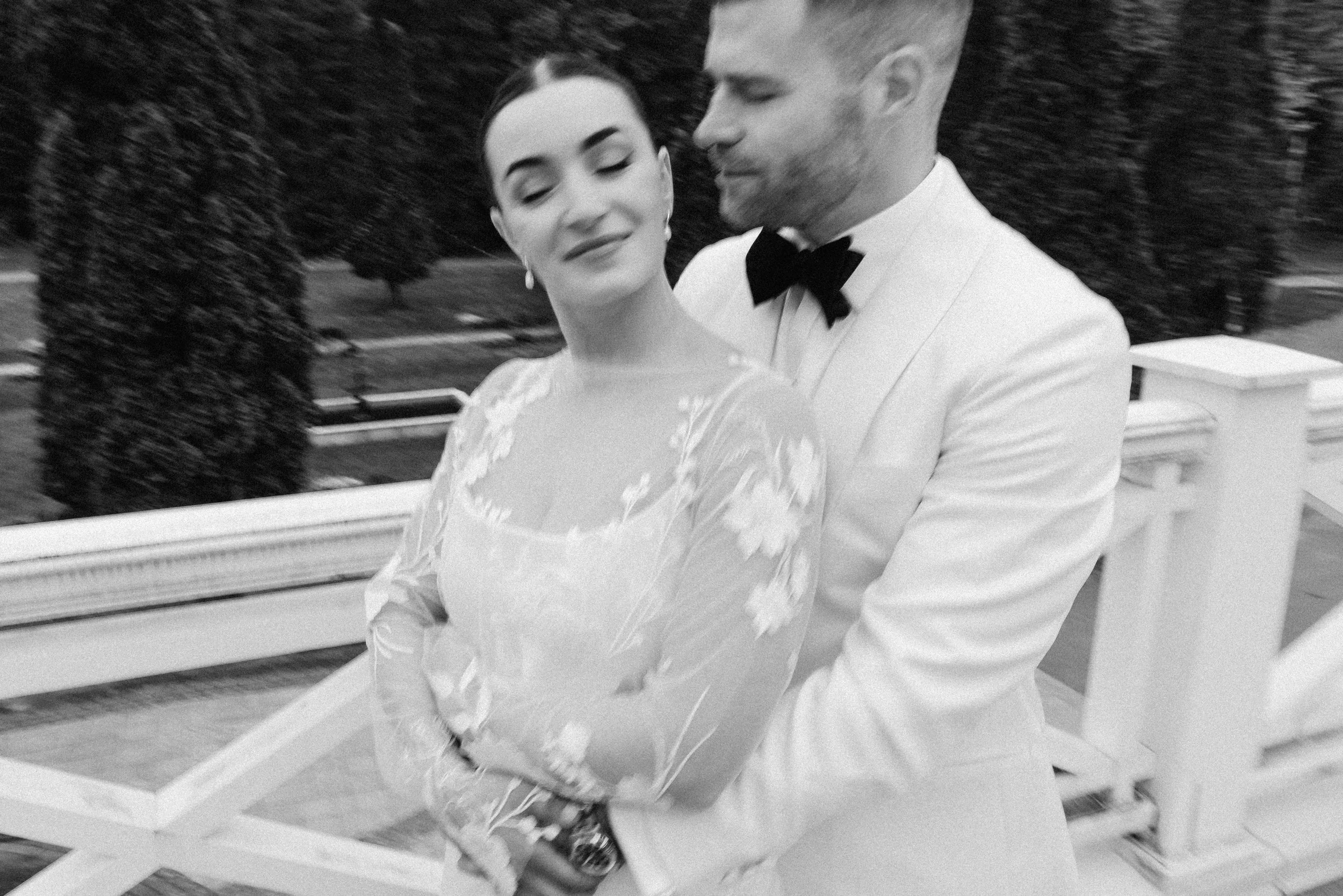 A black-and-white photograph of a couple dancing on a wedding dance floor. The woman has dark, short hair and is wearing earrings and a lace dress. The man has light-colored hair styled and is wearing a white tuxedo with a black bow tie. They are hol