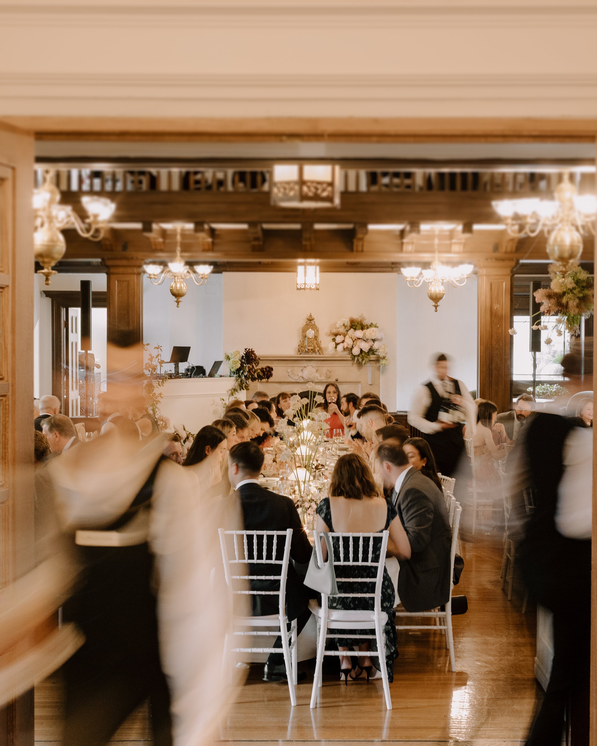 An elegant banquet hall filled with guests seated at long dining tables decorated with flowers. The room features wooden accents, chandeliers, and large windows letting in natural light.