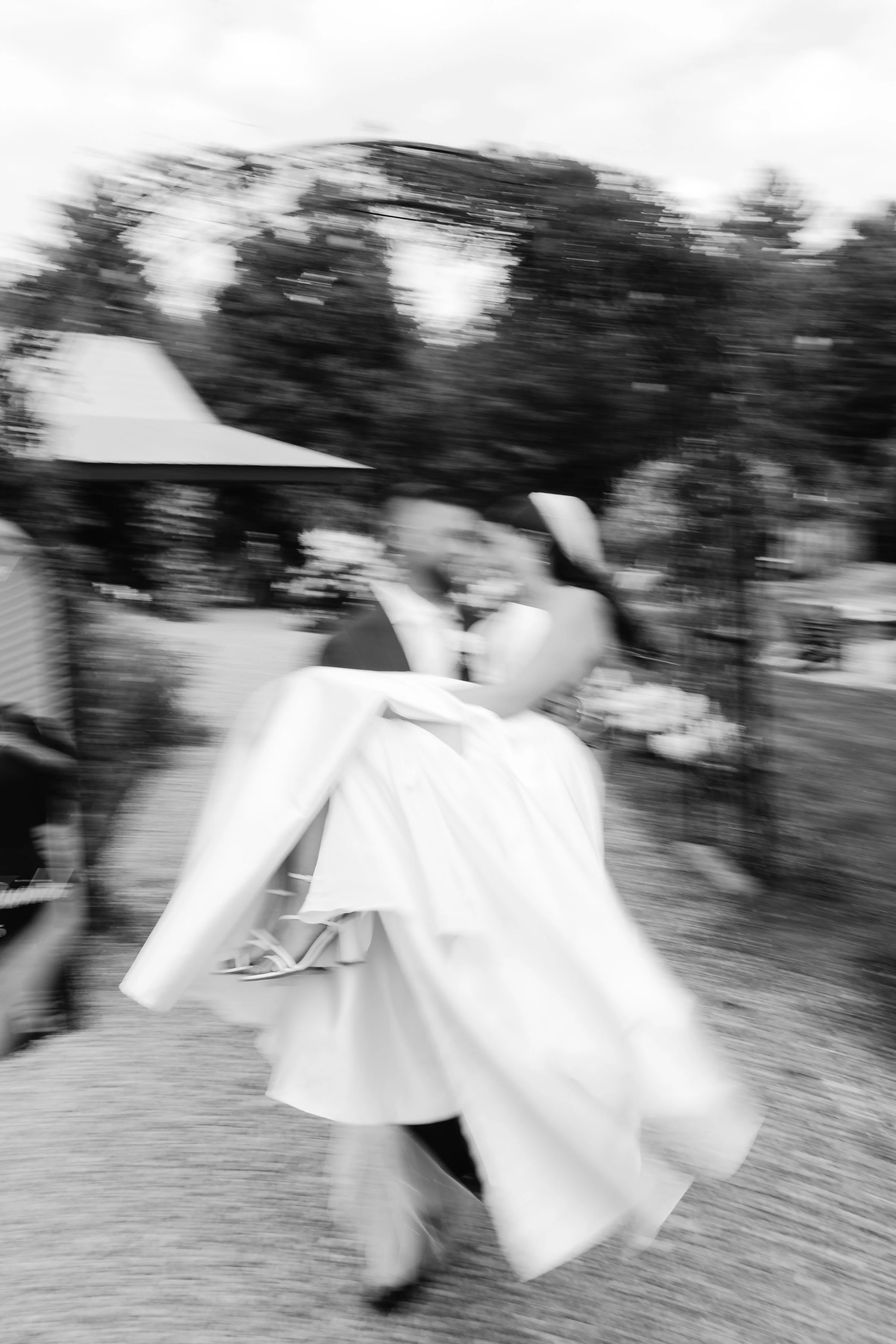 A person dressed in a long white gown and black shoes is walking in front of a backdrop of trees. The image is blurry and appears to be taken with motion blur.