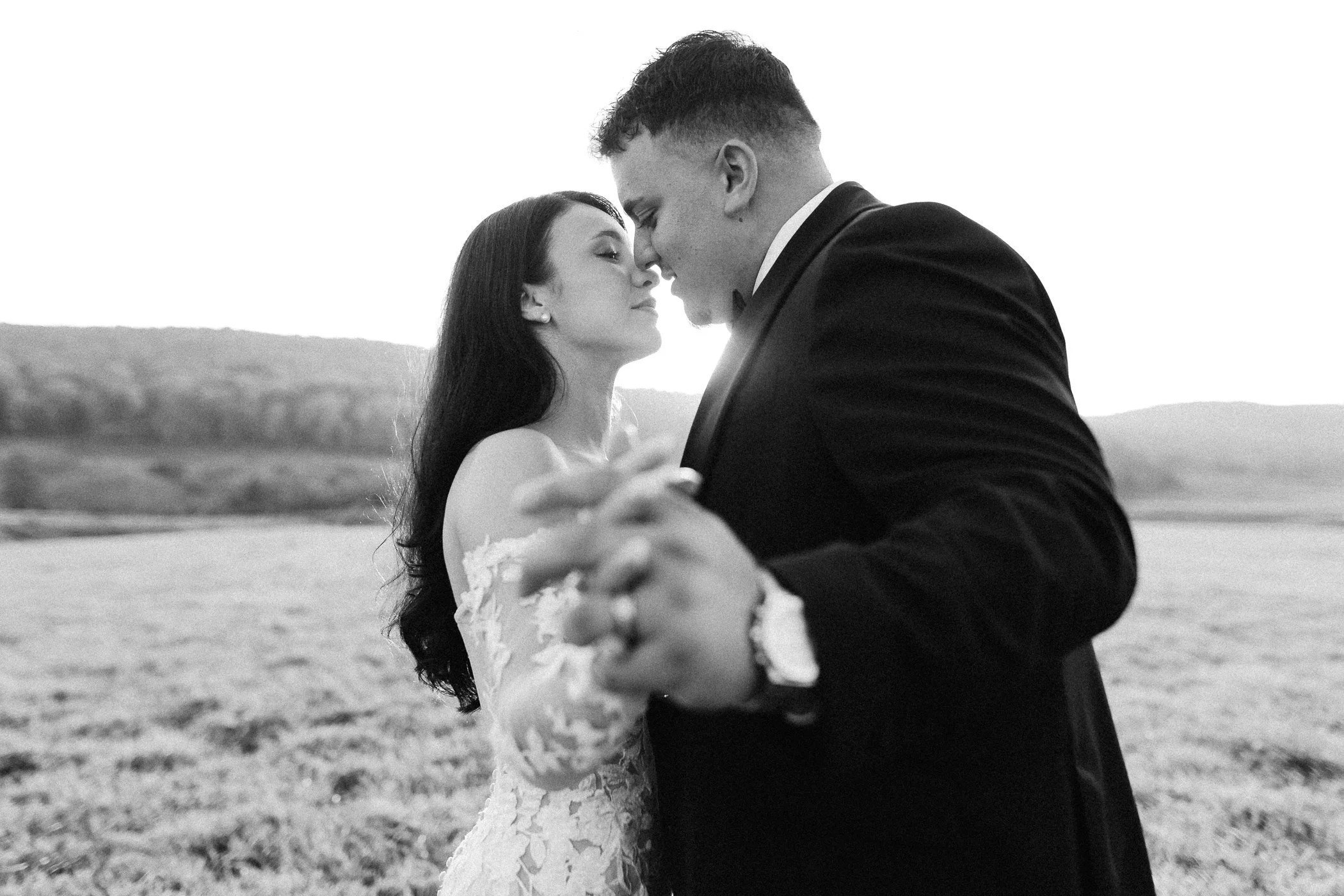 A black-and-white photo of a couple dancing outdoors, with hills in the background. The woman has long dark hair and is wearing a wedding dress, and the man is in a tuxedo. Their faces are close together as they dance.